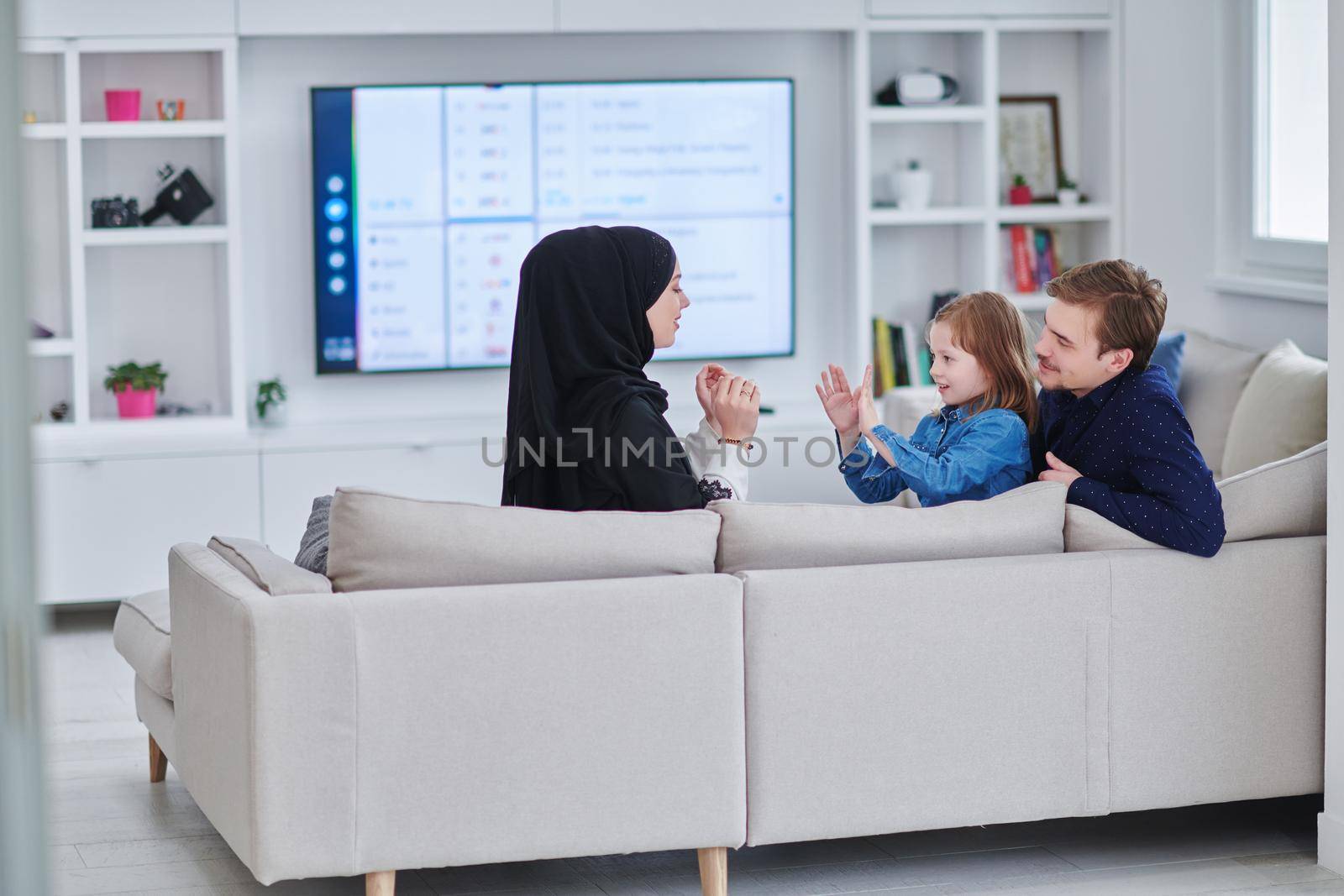 Happy Muslim family spending time together in modern home. Parents playing with daughter on the sofa during Ramadan. Traditional fashionable arabic clothes.