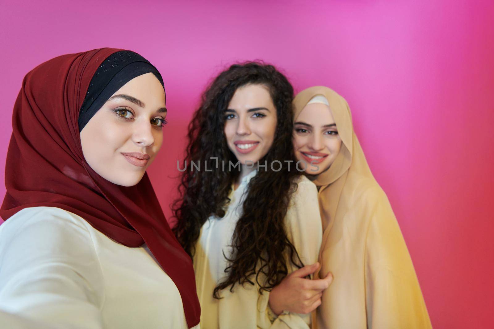 Muslim women taking selfie by mobile phone isolated on pink background. Group of pretty Arab girls in modern fashionable dress using technology