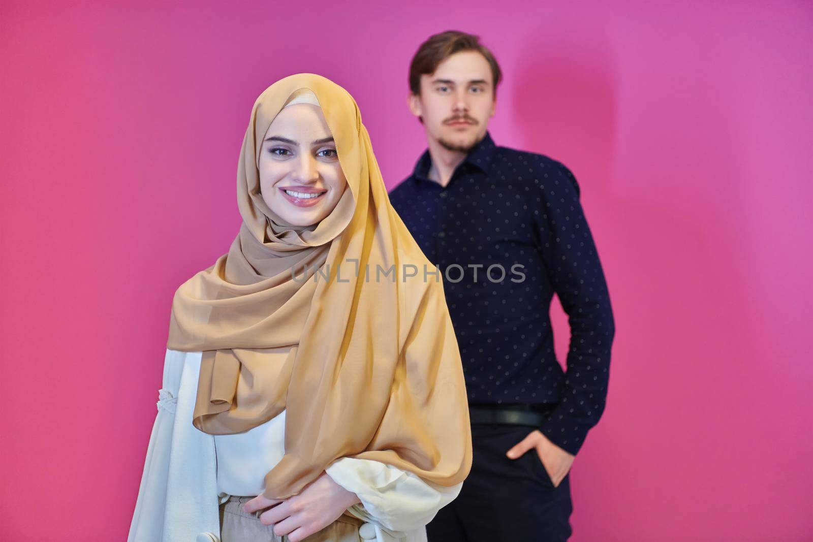 Portrait of happy young muslim couple standing isolated on pink background. Girl wearing modern hijab representing Ramadan concept