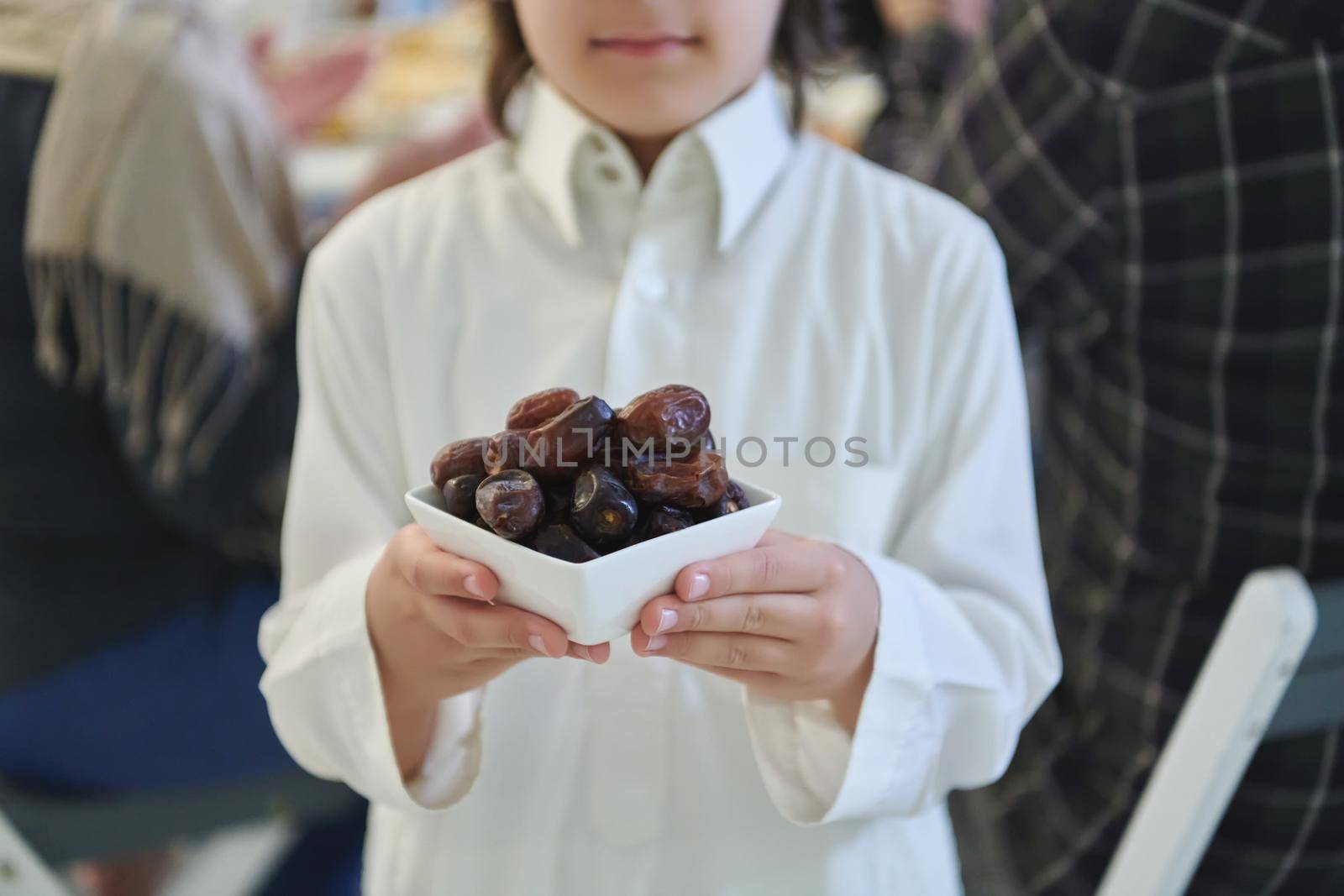 Arabian kid in the traditional clothes during iftar. Portrait of happy young muslim boy holding bowl of dates while rest of the family are eating