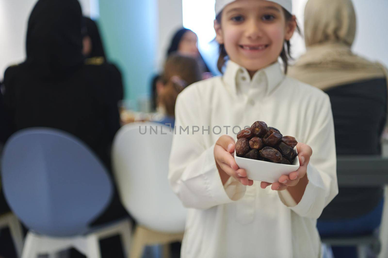 Arabian kid in the traditional clothes during iftar. Portrait of happy young muslim boy holding bowl of dates while rest of the family are eating