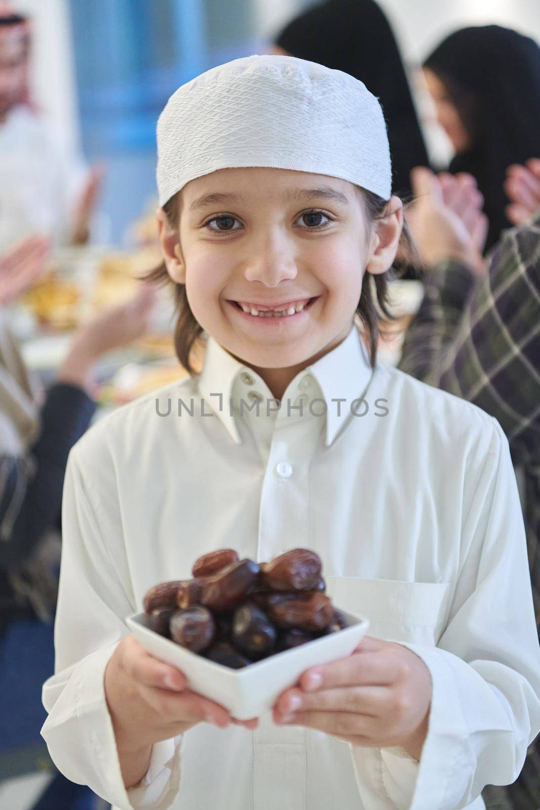 Arabian kid in the traditional clothes during iftar. Portrait of happy young muslim boy holding bowl of dates while rest of the family are eating