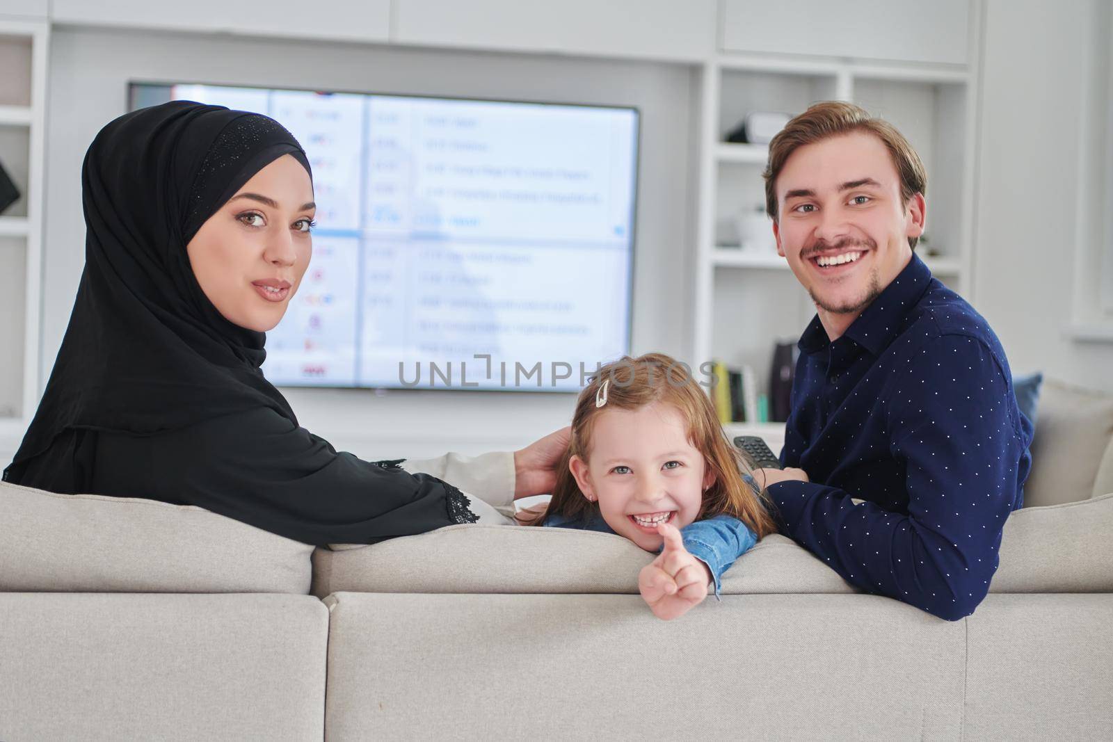 Happy Muslim family spending time together in modern home. Parents playing with daughter on the sofa during Ramadan. Traditional fashionable arabic clothes.