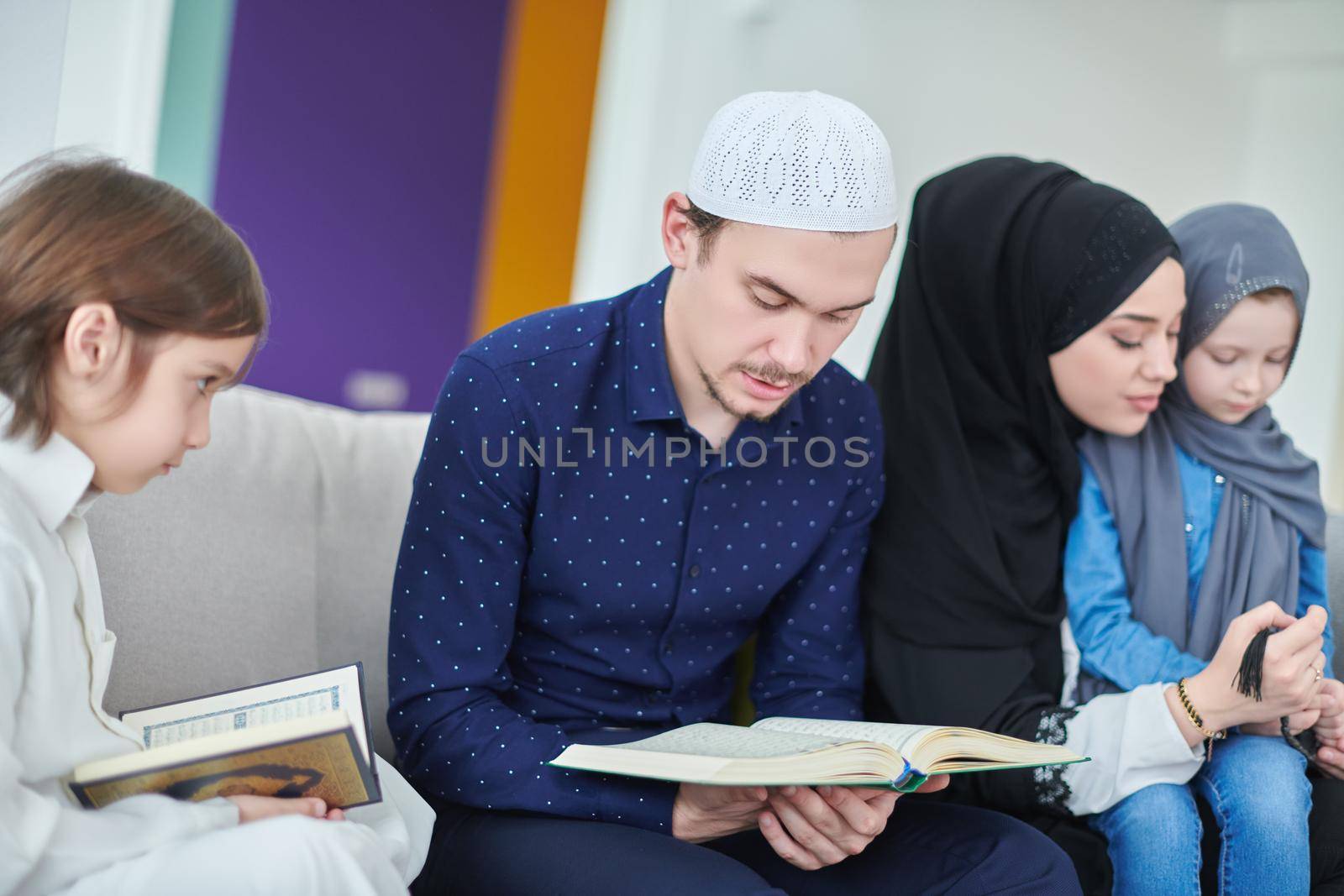 Young muslim family reading Quran during Ramadan. Parents and kids worshiping to God, in islamic clothes at modern home