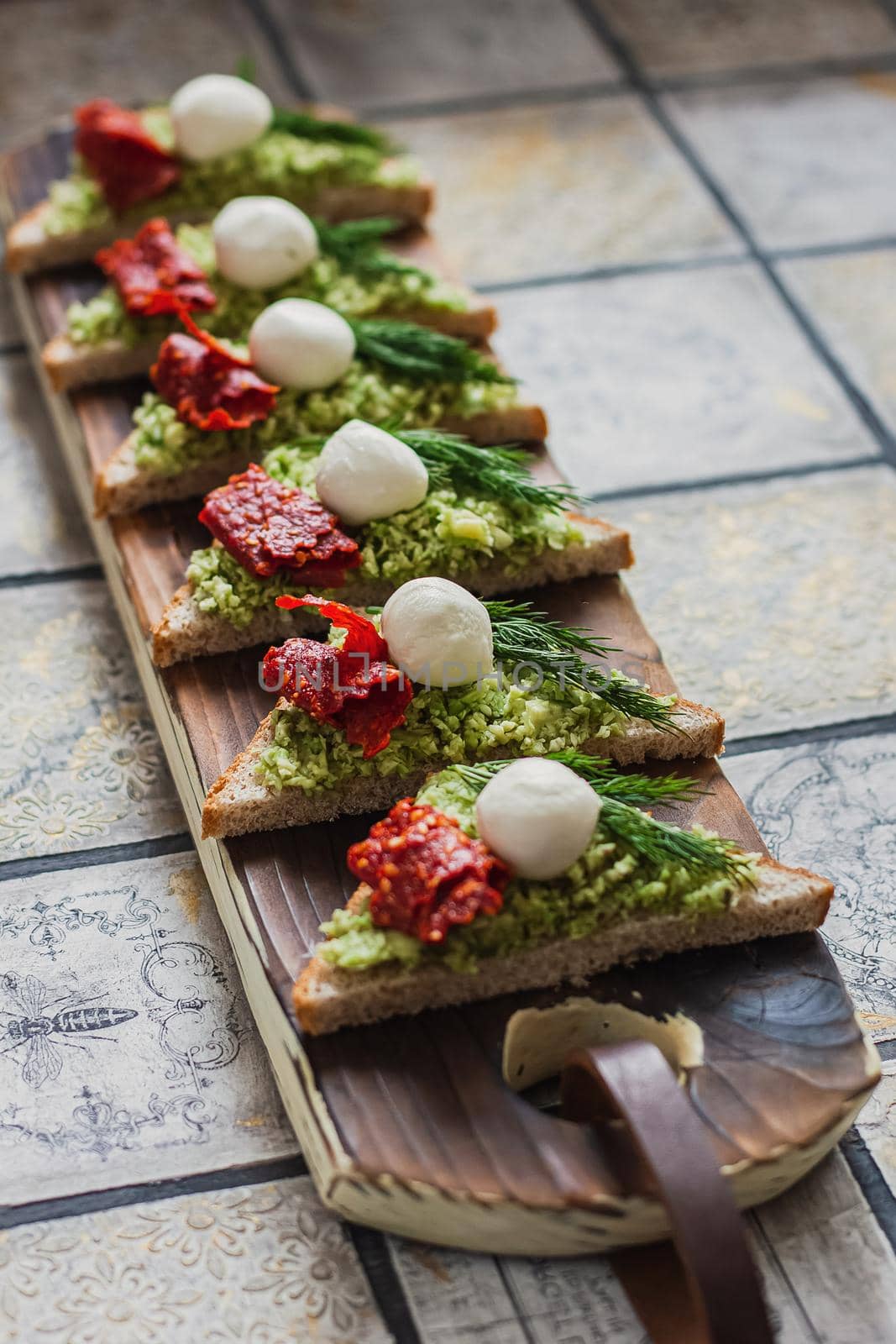 Mini triangle canapes bruschetta with avocado, mozarella and dried tomatos on the rustic board on the vintage tiled background by mmp1206