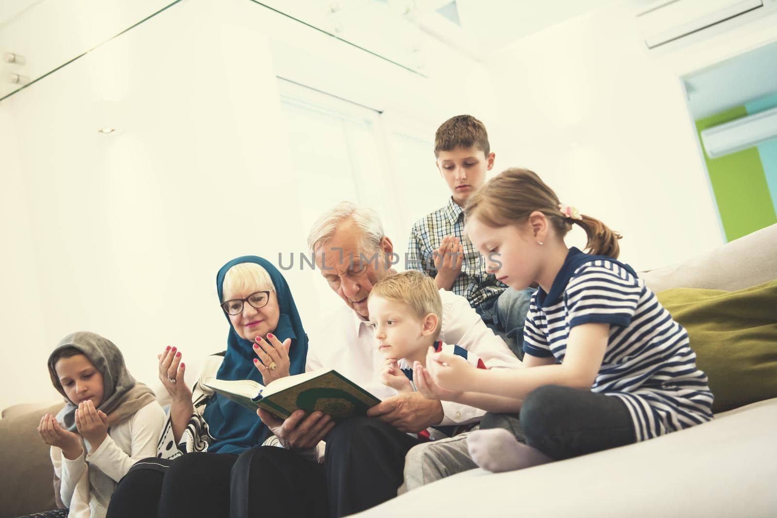 modern muslim family grandparents with grandchildren reading Quran and praying together on the sofa before iftar dinner during a ramadan feast at home