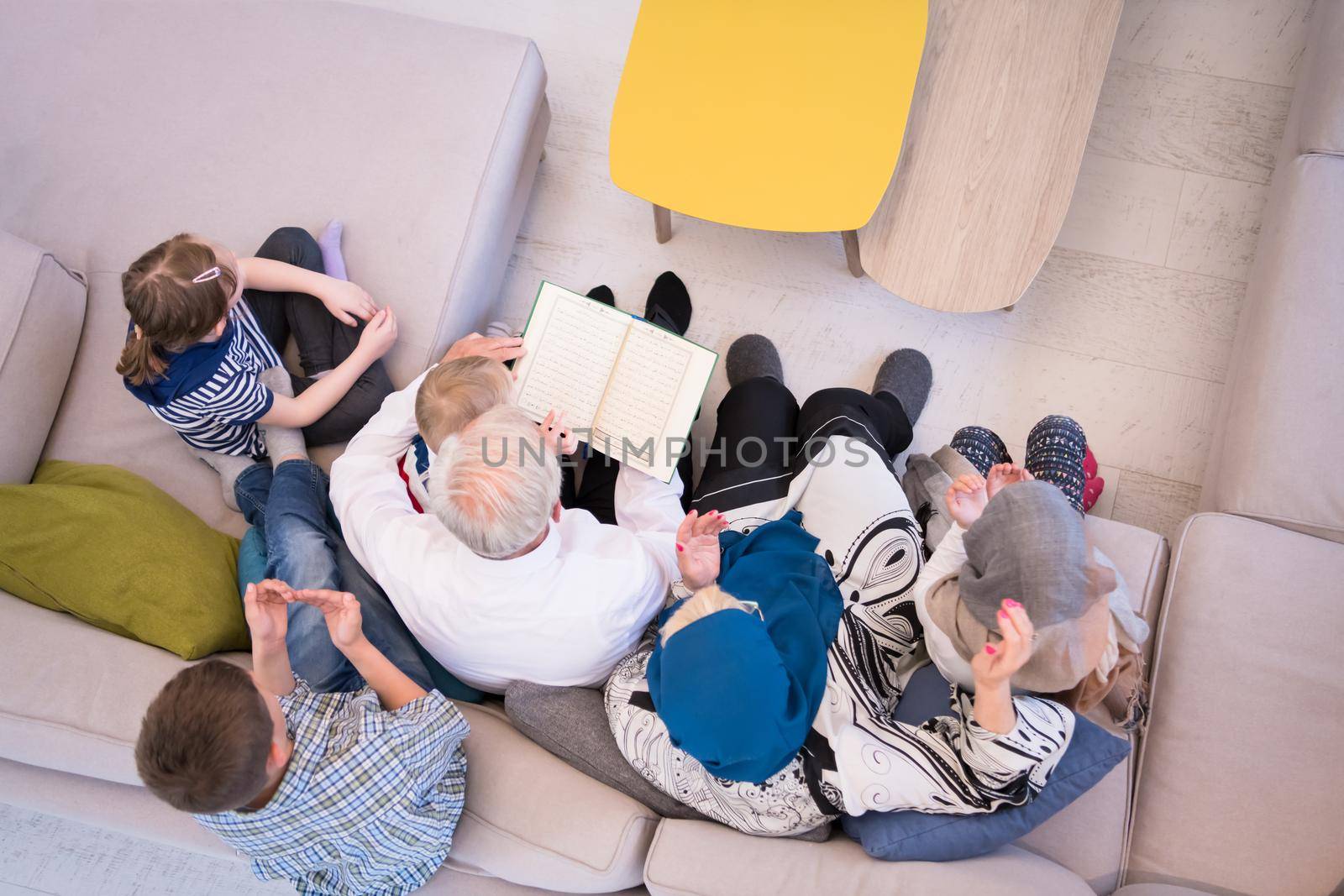 top view of modern muslim family grandparents with grandchildren reading Quran and praying together on the sofa before iftar dinner during a ramadan feast at home