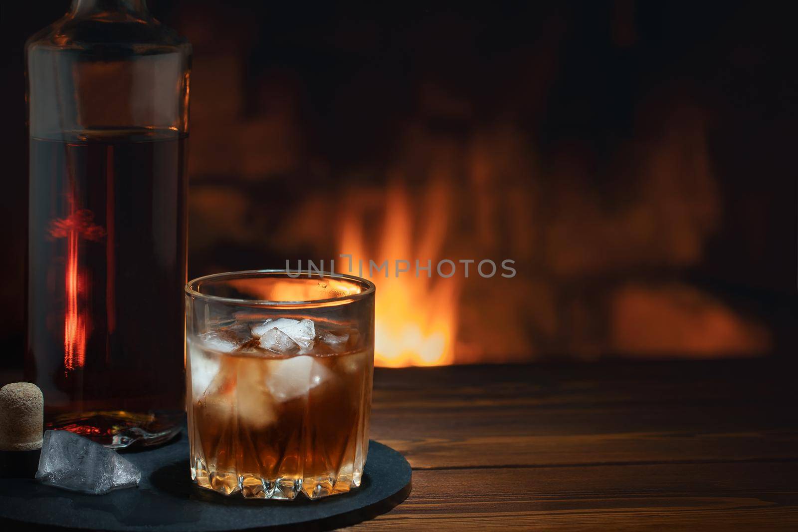 Bottle and glass of whiskey with ice on the table near the burning fireplace. Rest and relaxation concept.