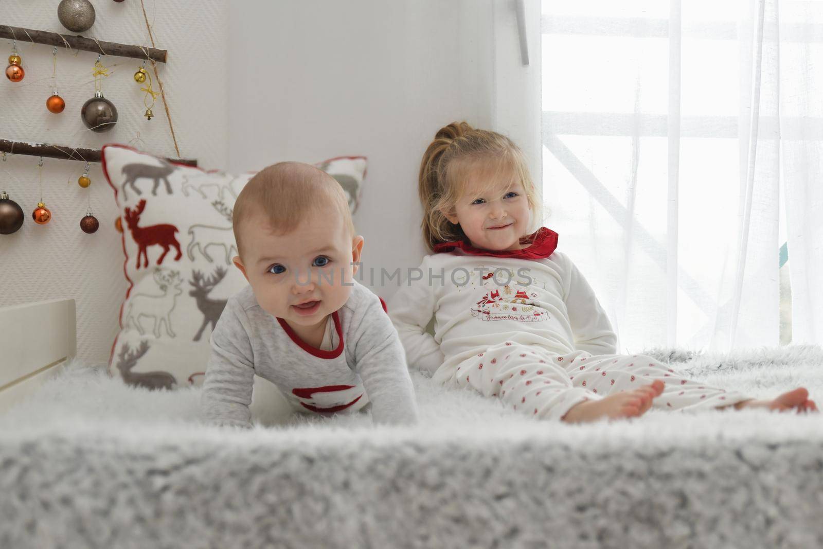 Brother and sister in Christmas pajamas are sitting on the bed with decorations.