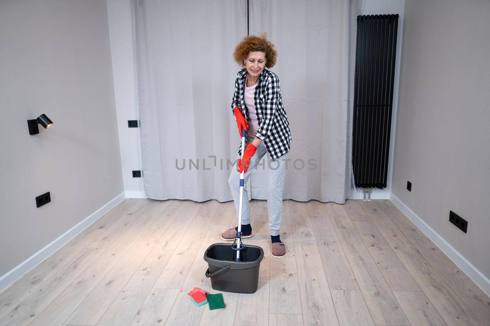 People, housework and housekeeping concept. Happy senior woman in protective gloves cleans floor and dances at home in empty apartment before moving to new home. Excited woman enjoying housekeeping.