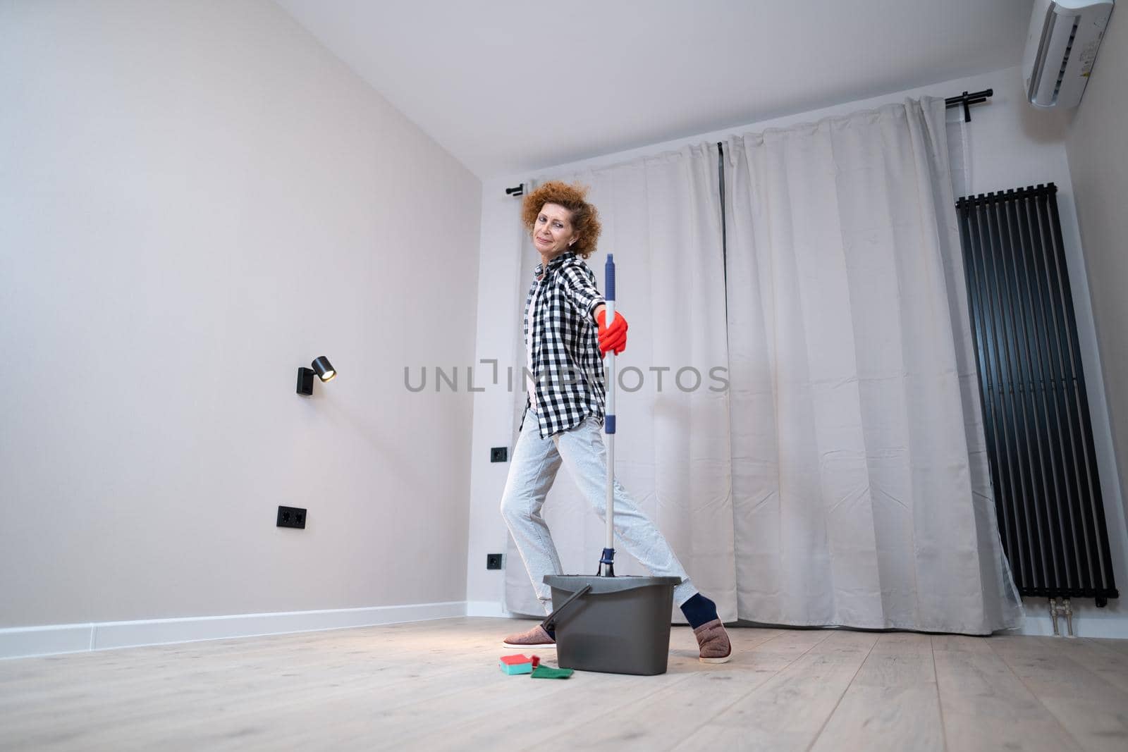 Jolly excited mature woman enjoying cleaning house, she dancing while washing floor. Happy elderly woman enjoying cleaning floors before moving to new apartment. Housework and housekeeping concept by Tomashevska