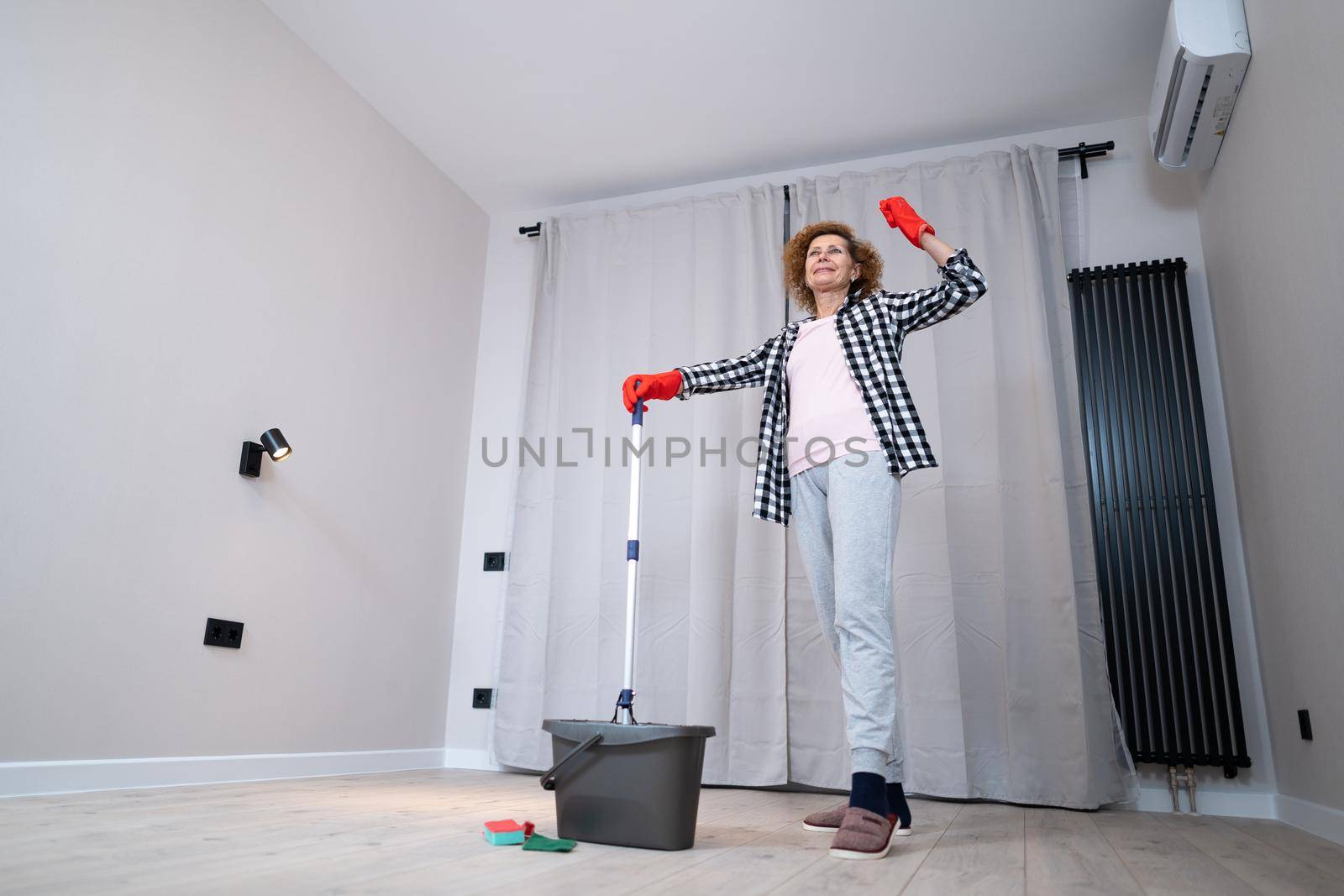 Happy mature woman mops the floor and dances and sings in a new unfurnished apartment before moving to a new house. Housewife enjoying domestic chores, doing home cleanup creatively by Tomashevska