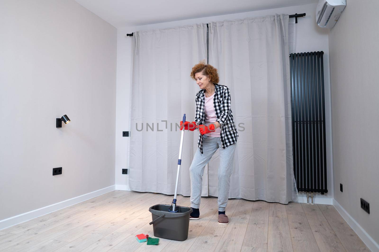 Happy mature woman mops the floor and dances and sings in a new unfurnished apartment before moving to a new house. Housewife enjoying domestic chores, doing home cleanup creatively by Tomashevska