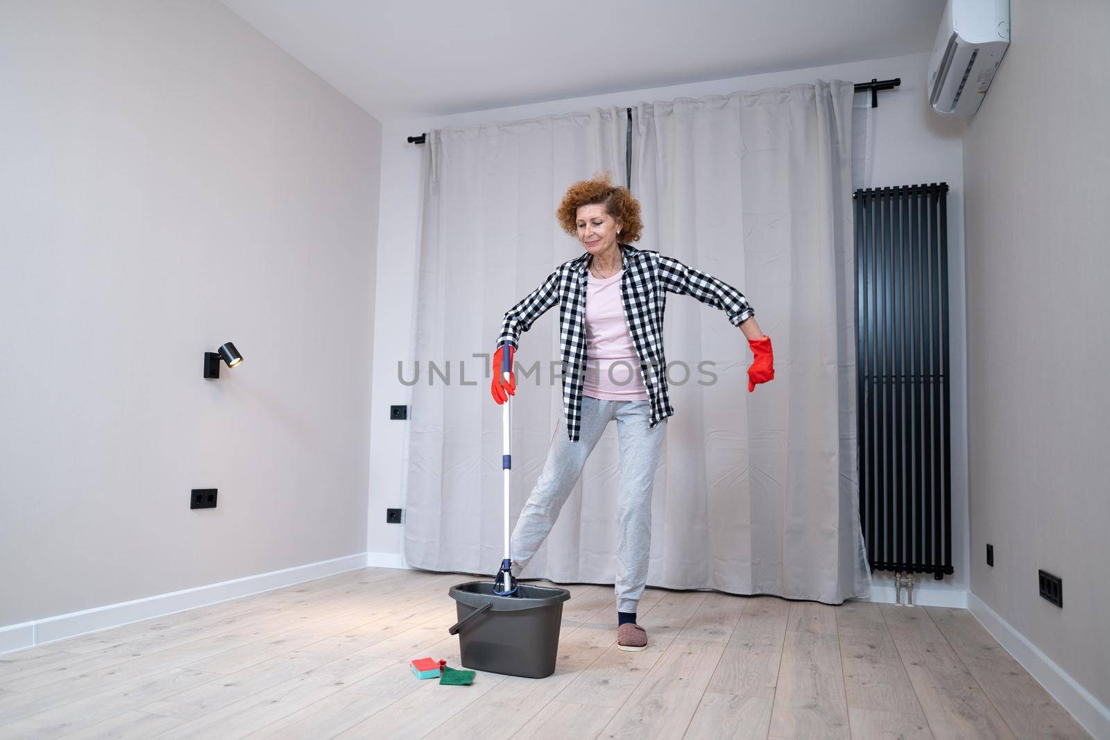 Happy mature woman mops the floor and dances and sings in a new unfurnished apartment before moving to a new house. Housewife enjoying domestic chores, doing home cleanup creatively.