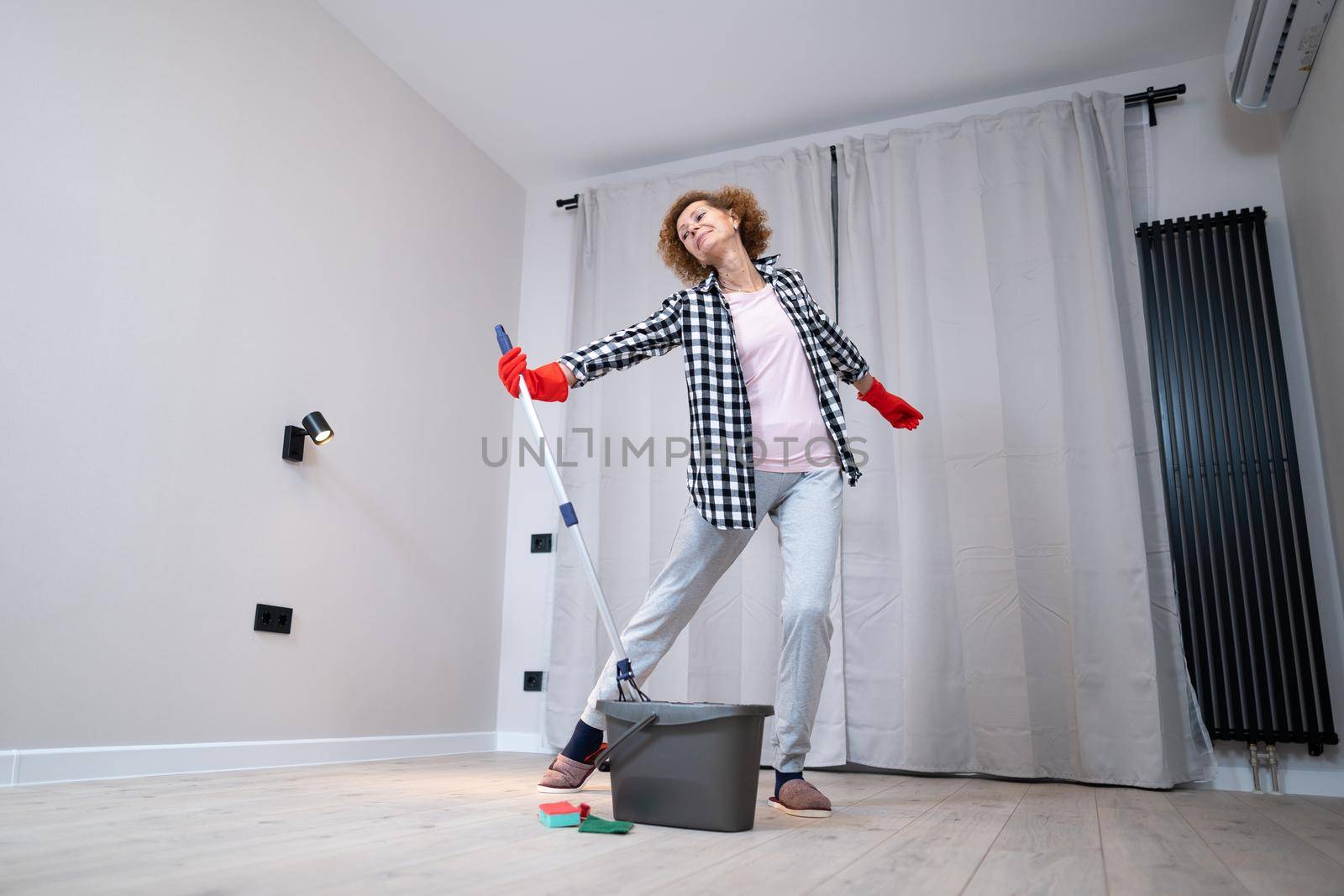 People, housework and housekeeping concept. Happy senior woman in protective gloves cleans floor and dances at home in empty apartment before moving to new home. Excited woman enjoying housekeeping.