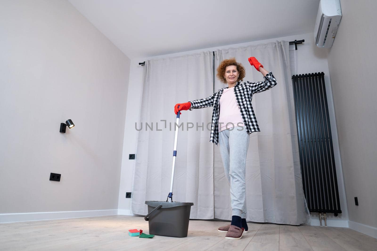 People, housework and housekeeping concept. Happy senior woman in protective gloves cleans floor and dances at home in empty apartment before moving to new home. Excited woman enjoying housekeeping.