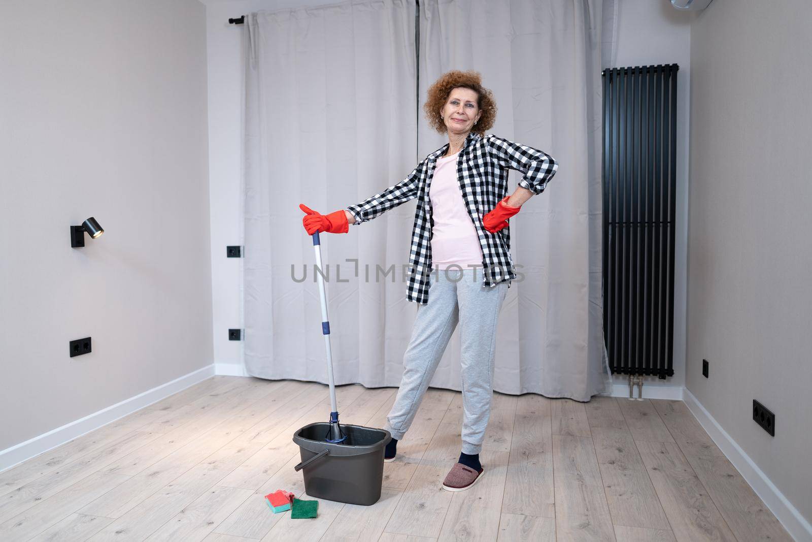 People, housework and housekeeping concept. Happy senior woman in protective gloves cleans floor and dances at home in empty apartment before moving to new home. Excited woman enjoying housekeeping by Tomashevska