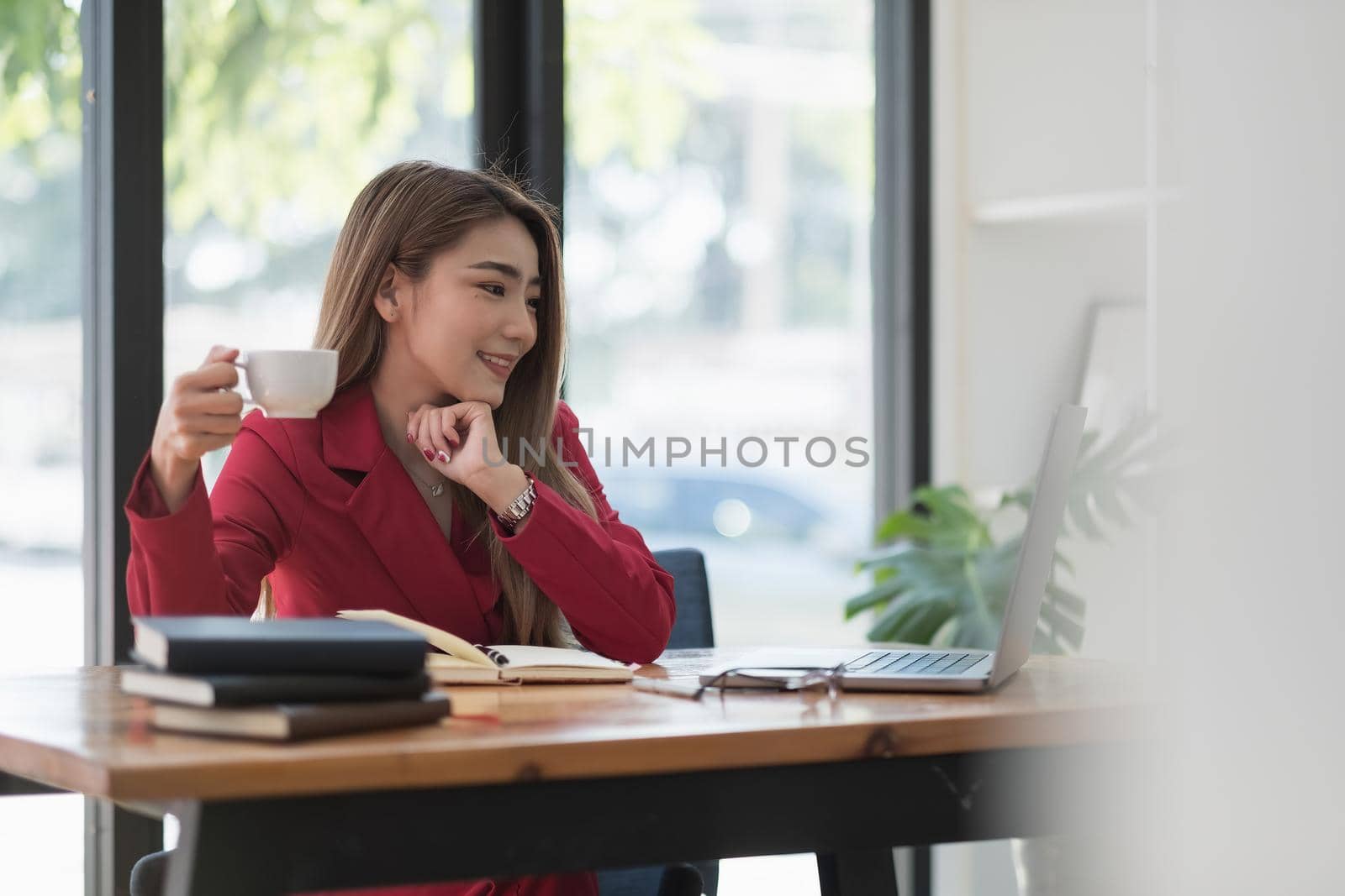 Charming asian businesswoman sitting working with laptop in office. Smiling hand with cup of coffee in morning.