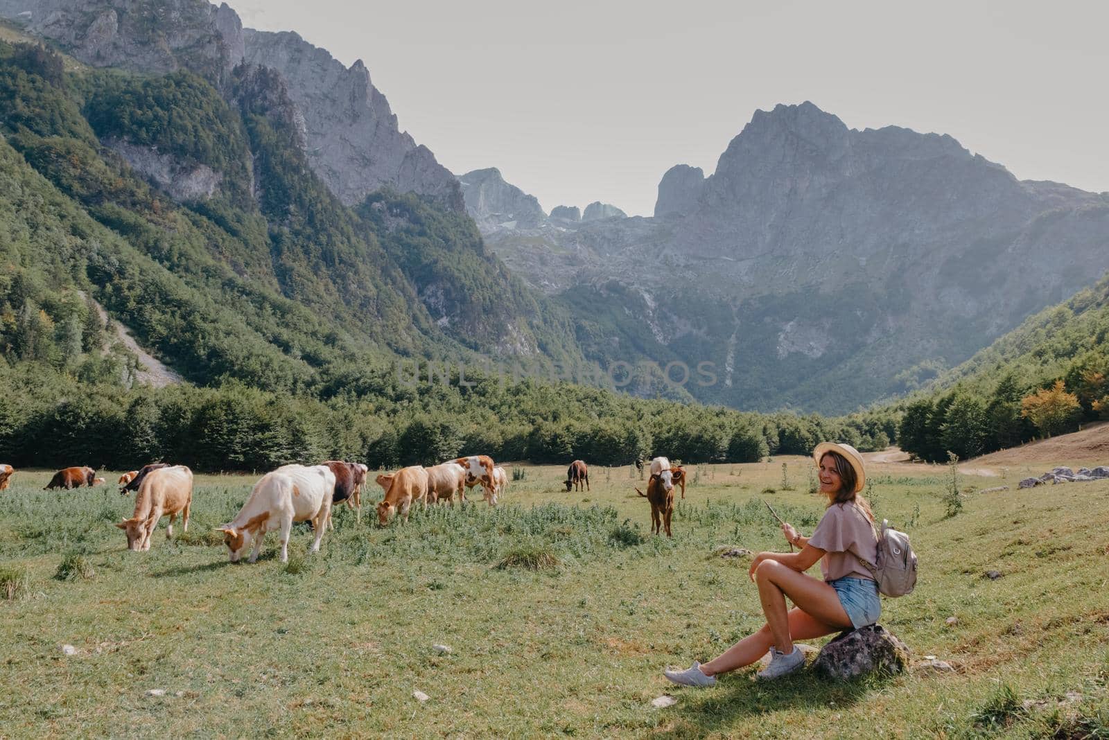 Travel, Lifestyle Concept. Beautiful woman enjoys views of the alpine village in the Alps mountains. Young woman sitting and relaxing on alpine mountain looking the cow eating a grass in the summer. Happy tourist girl traveling to Europe by Andrii_Ko