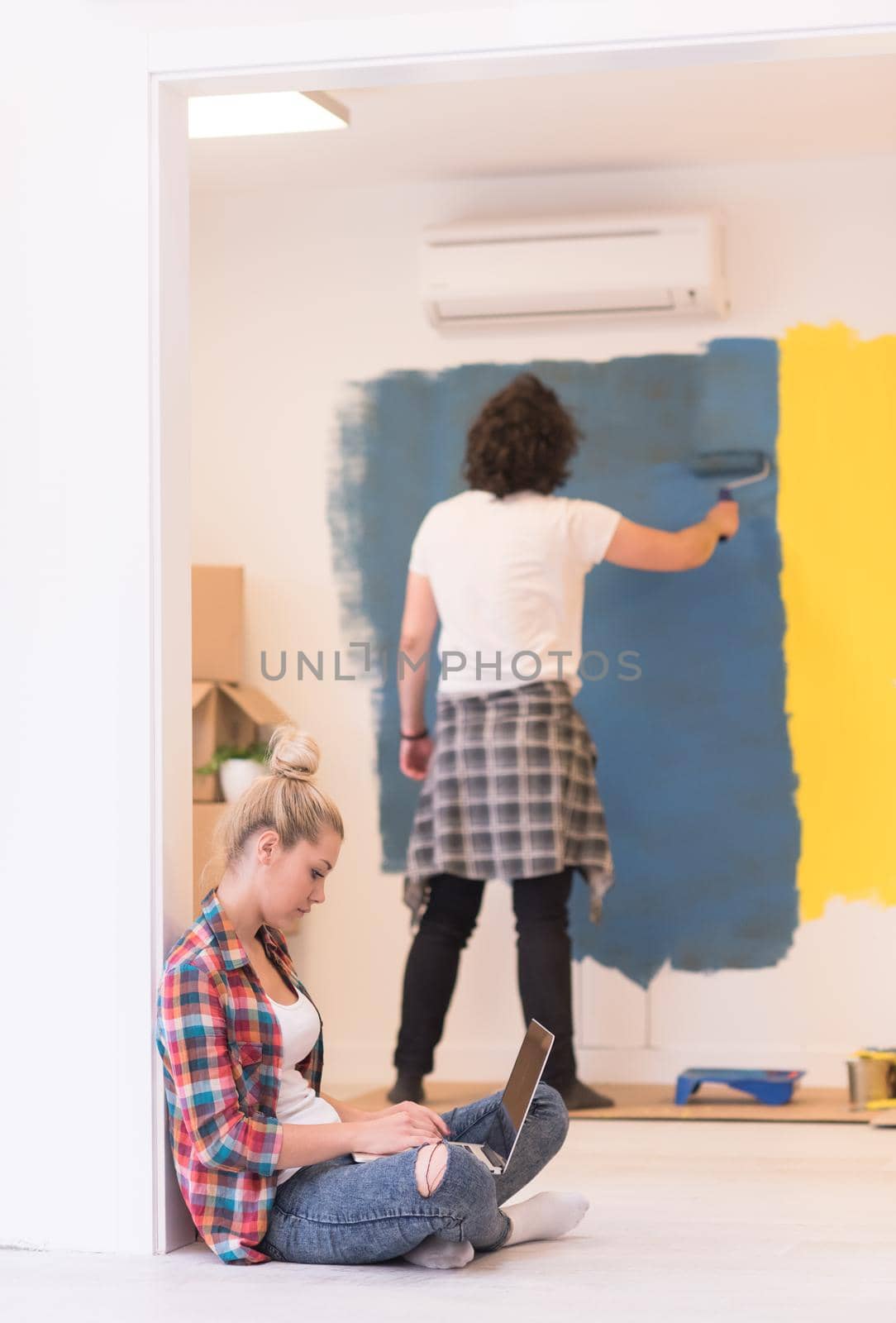 Happy couple doing home renovations, the man is painting the room and the woman is relaxing on the floor and connecting with a laptop