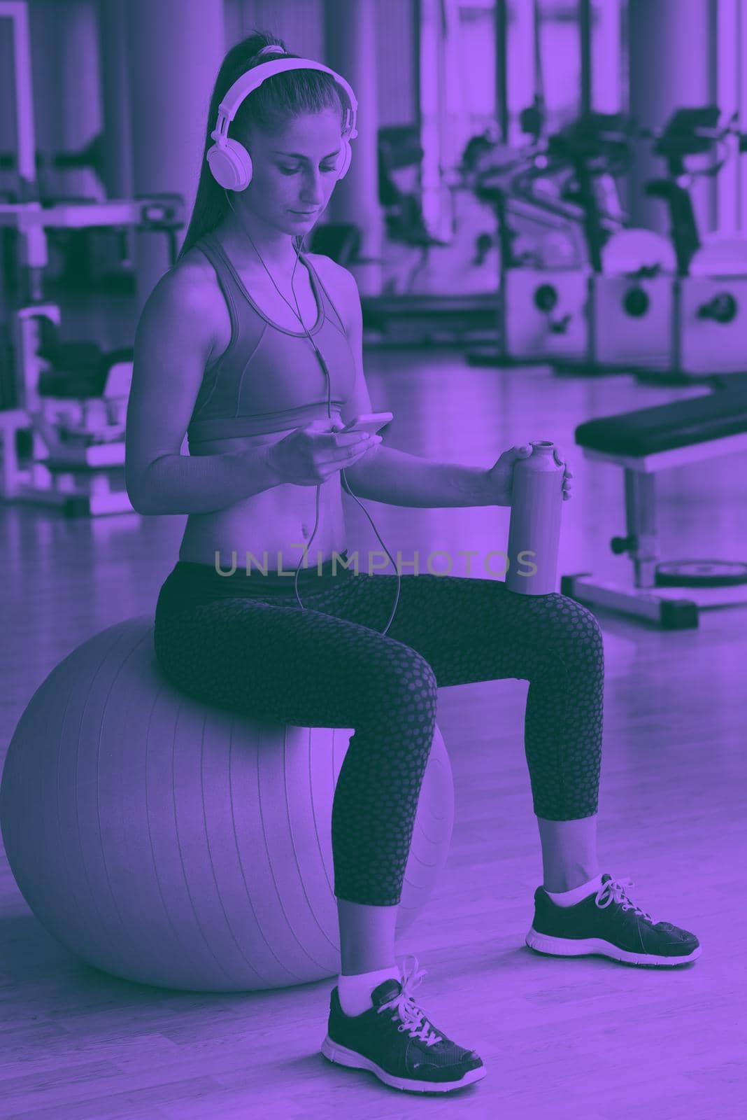 young healthy woman drinking water  in fitness gym while sitting on pilates ball and listening music on headphones from smartphone duo tone