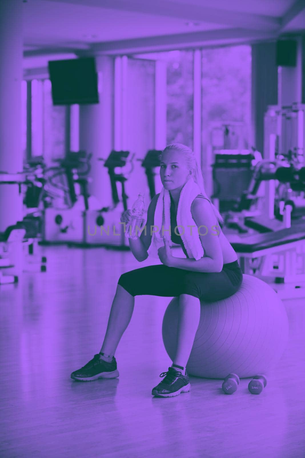 young healthy blonde woman drinking water  in fitness gym while sitting on pilates ball duo tone