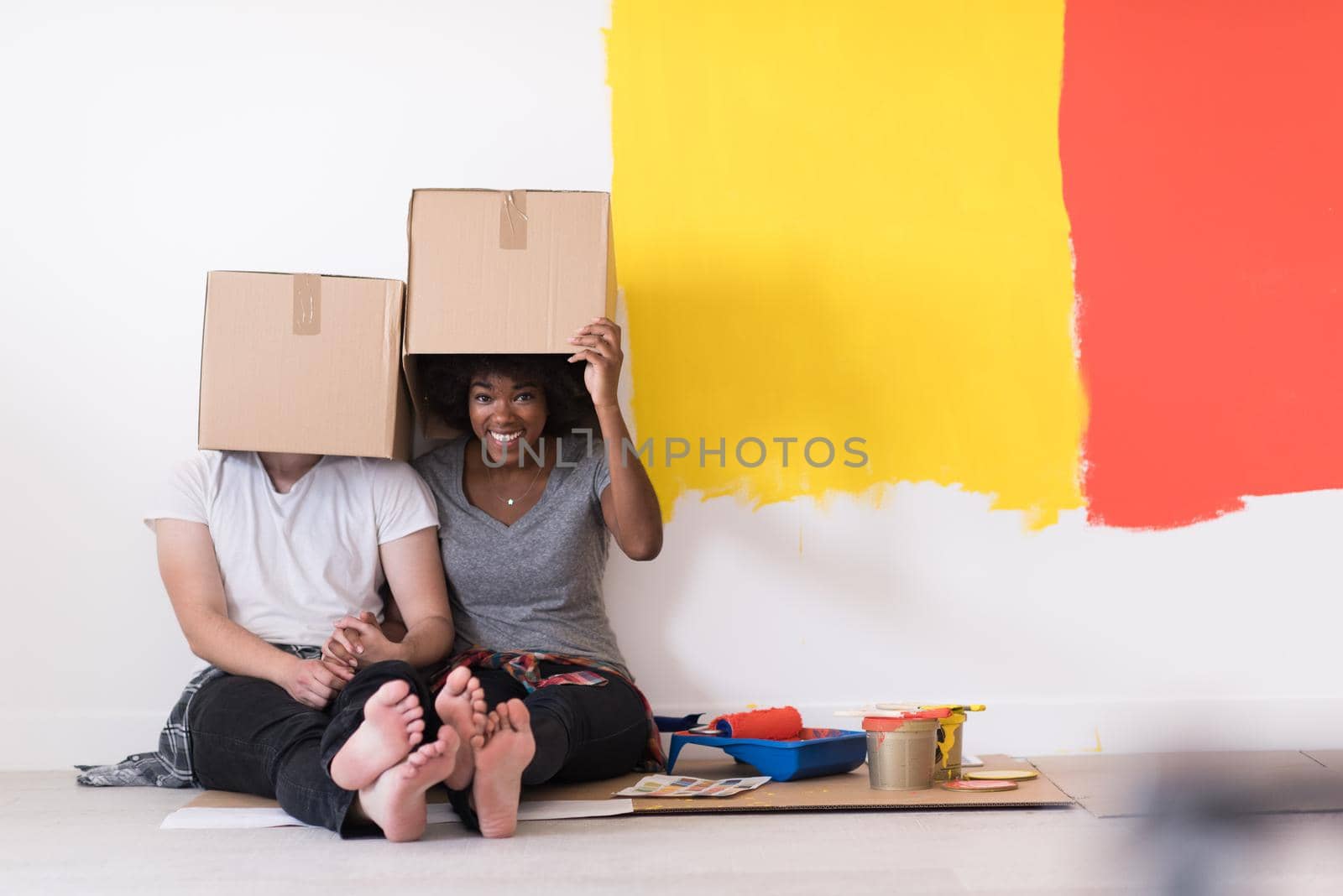 Happy young multiethnic couple relaxing and playing with cardboard boxes after painting a room in their new house on the floor