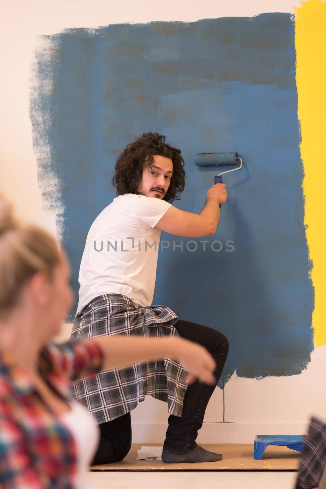 Happy couple doing home renovations, the man is painting the room and the woman is relaxing on the floor and connecting with a laptop