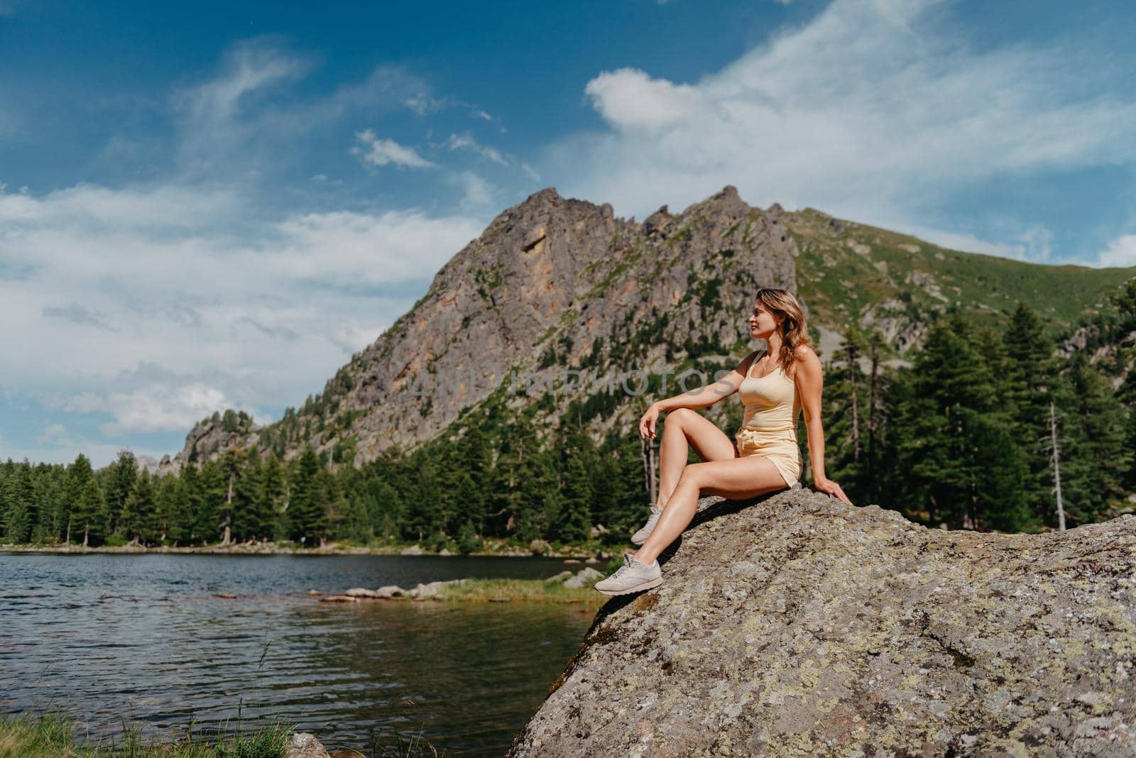 Tourist girl enjoys the magical view of the lake, coniferous forest and magical view sitting on big stone on the shore of a turquoise lake in the mountains. Hiking in the Natural Park. Cute girl tourist sitting on a large stone by the lake. The water is clear, stones are visible under the water. by Andrii_Ko
