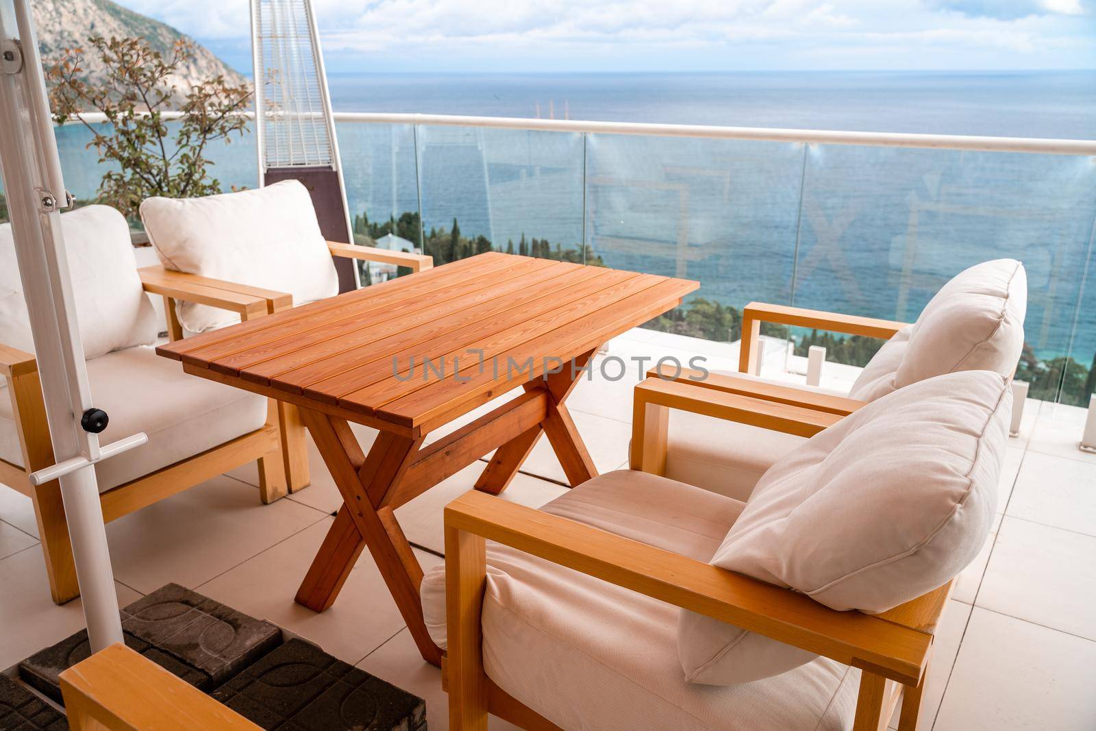 Restaurant with sea view. You can see rocks in the sea. Wooden tables and chairs with white cushions. Overcast, cloudy weather. by Matiunina