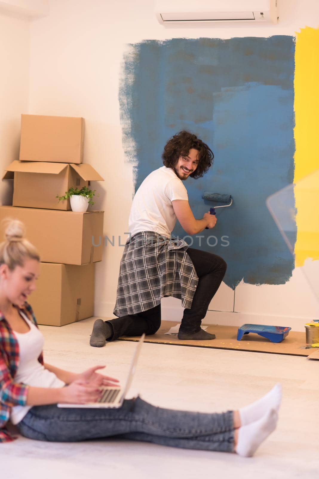 Happy couple doing home renovations, the man is painting the room and the woman is relaxing on the floor and connecting with a laptop