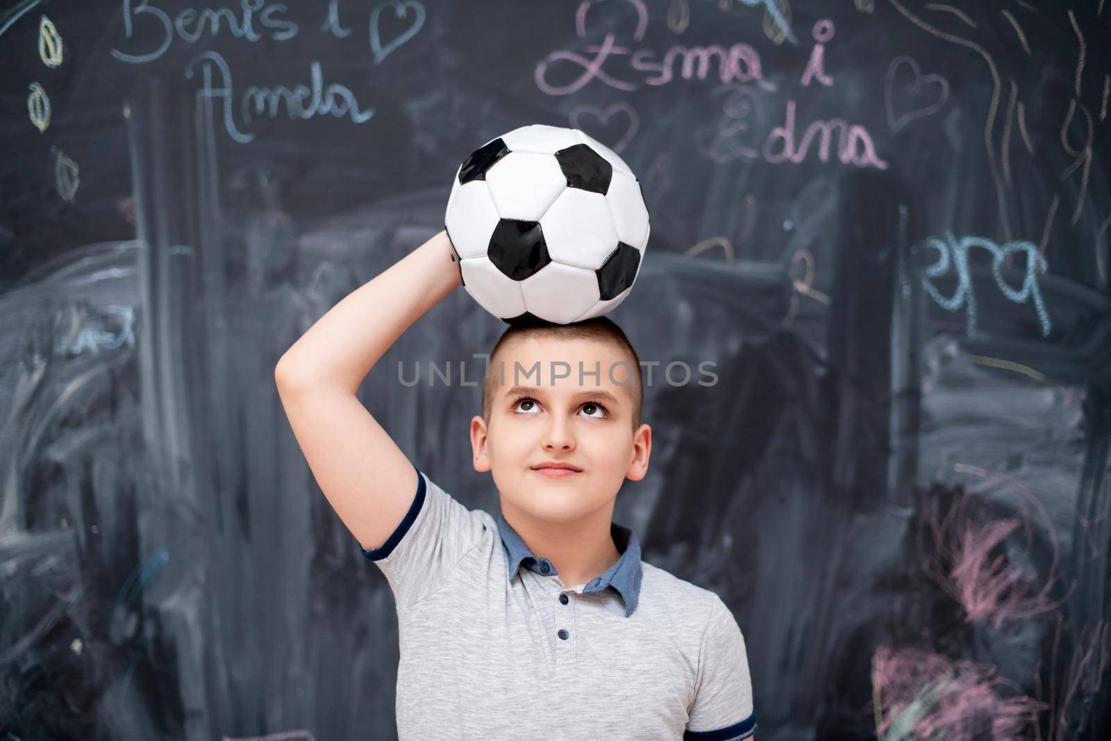 portrait of happy cute boy having fun holding a soccer ball on his head while standing in front of black chalkboard