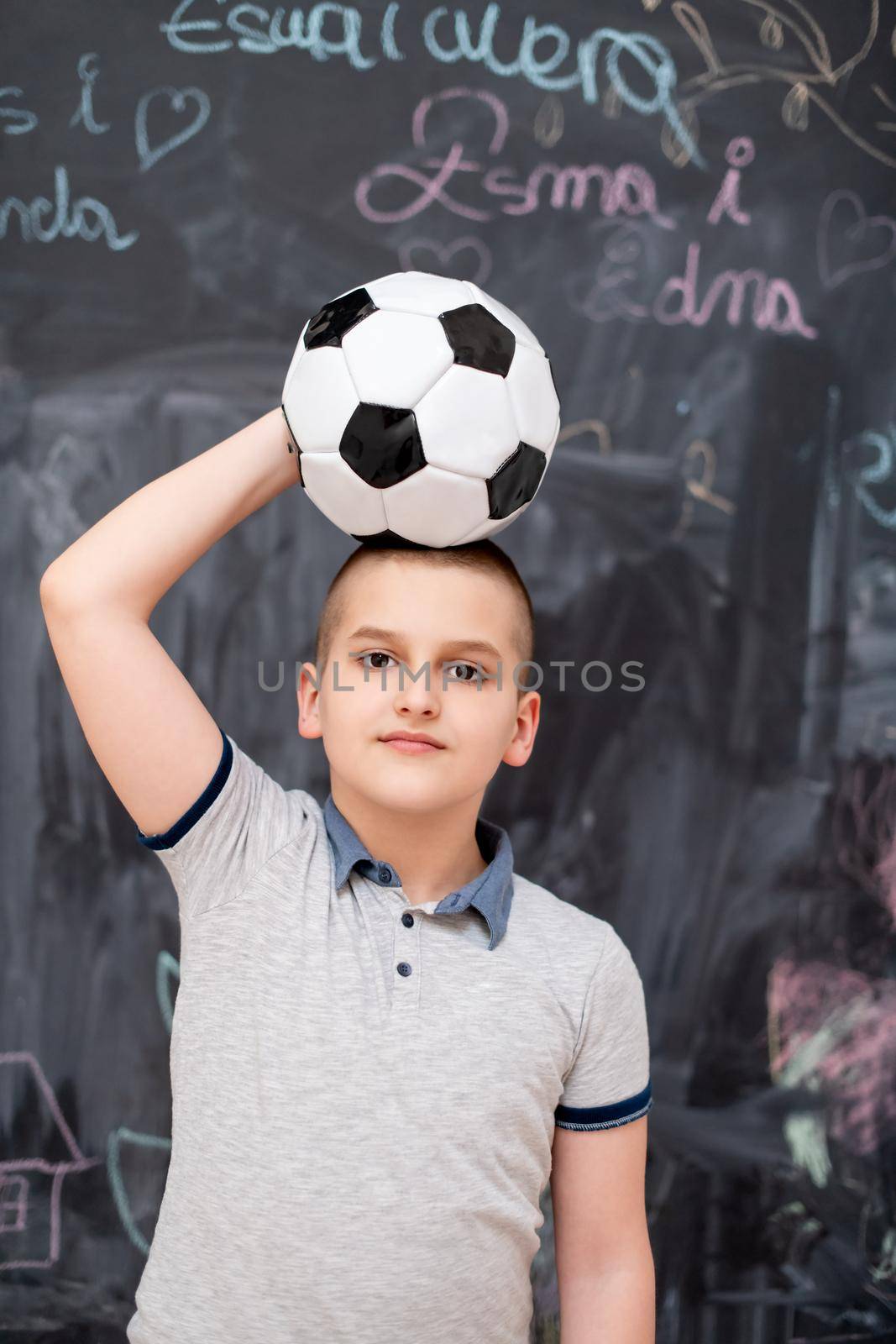 portrait of happy cute boy having fun holding a soccer ball on his head while standing in front of black chalkboard