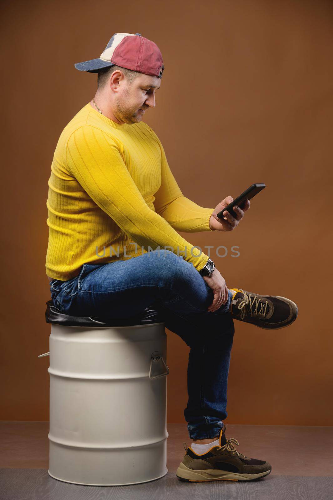 A plump man in casual clothes and a cap sits on a metal barrel with a phone in his hand. Writes SMS or checks mail. Studio portrait by yanik88