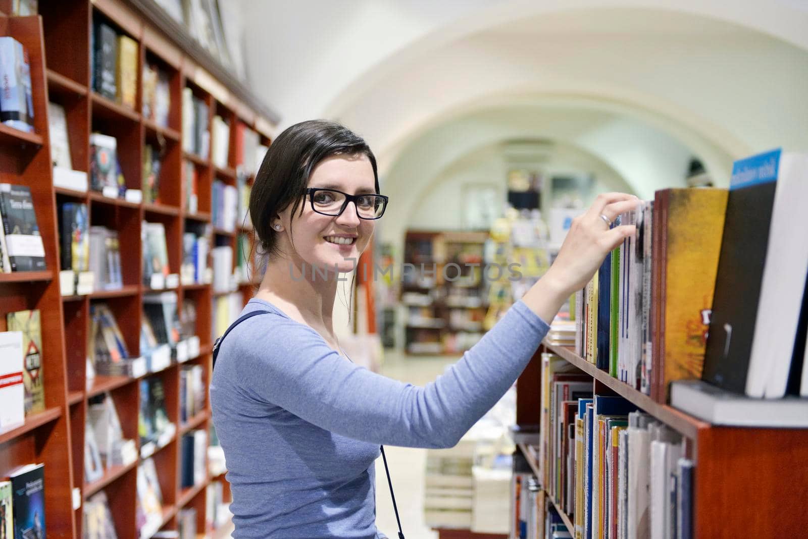 Pretty female student standing at bookshelf in university library store shop  searching for a book