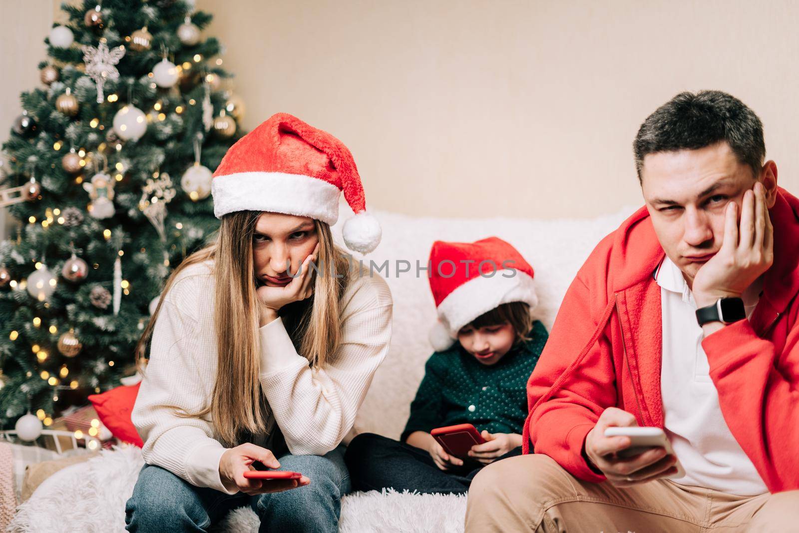 Portrait of depressed family with son playing cell phone during new year party. Tired mom and dad having a stressful time while kid using with mobile phone. Family dispute after Christmas celebration by Ostanina