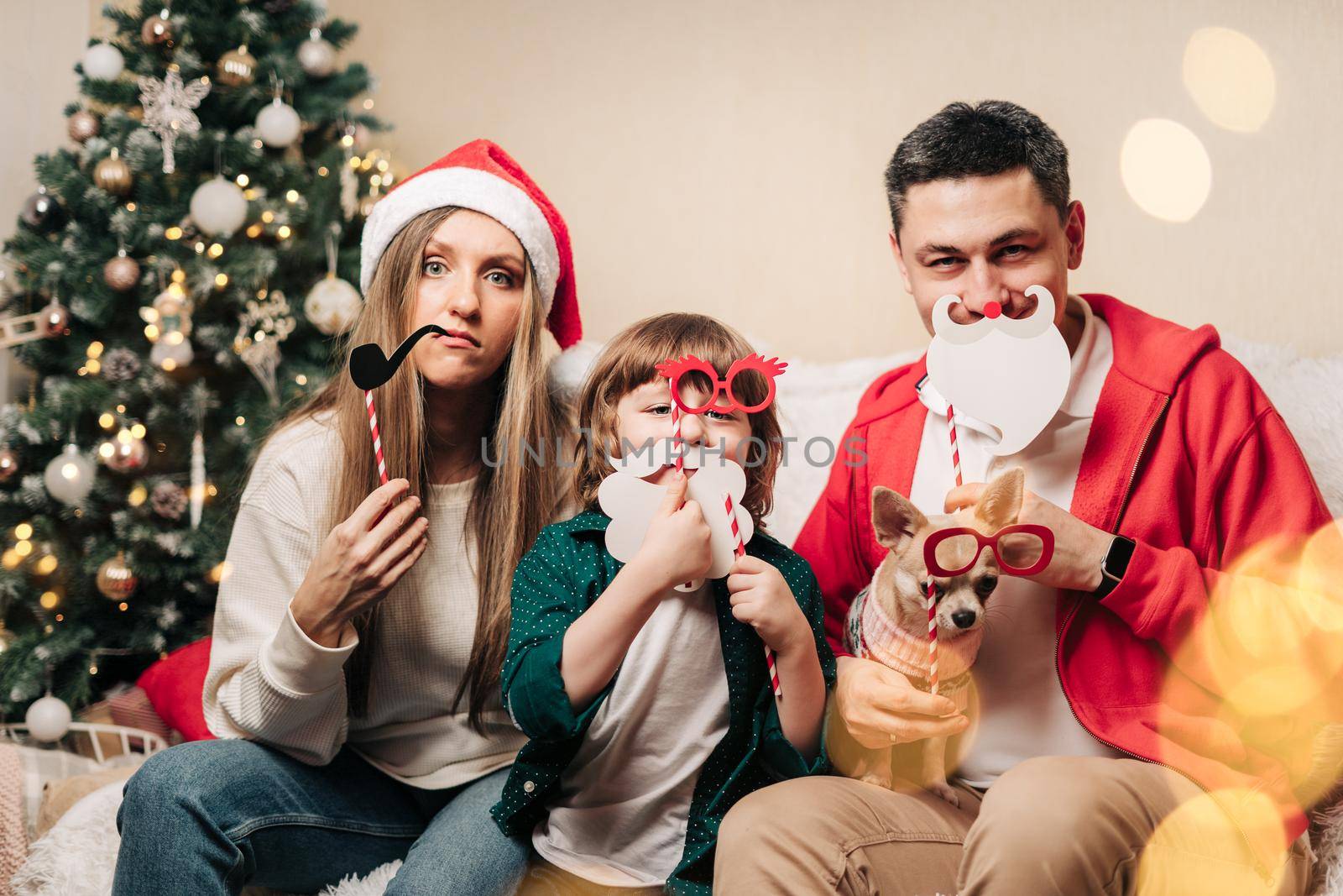 Portrait of happy family with kid son and puppy holding festive party props for photo booth. Mother in Santa hat, father, child boy and dog in sweater having fun on Christmas holidays at home by Ostanina