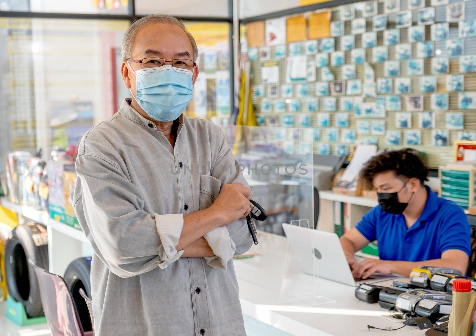 Senior man hold remote car key and arm-crossed with hygiene mask and stand in front of garage worker in counter service area. Automotive business and support management system concept for customer. by nrradmin