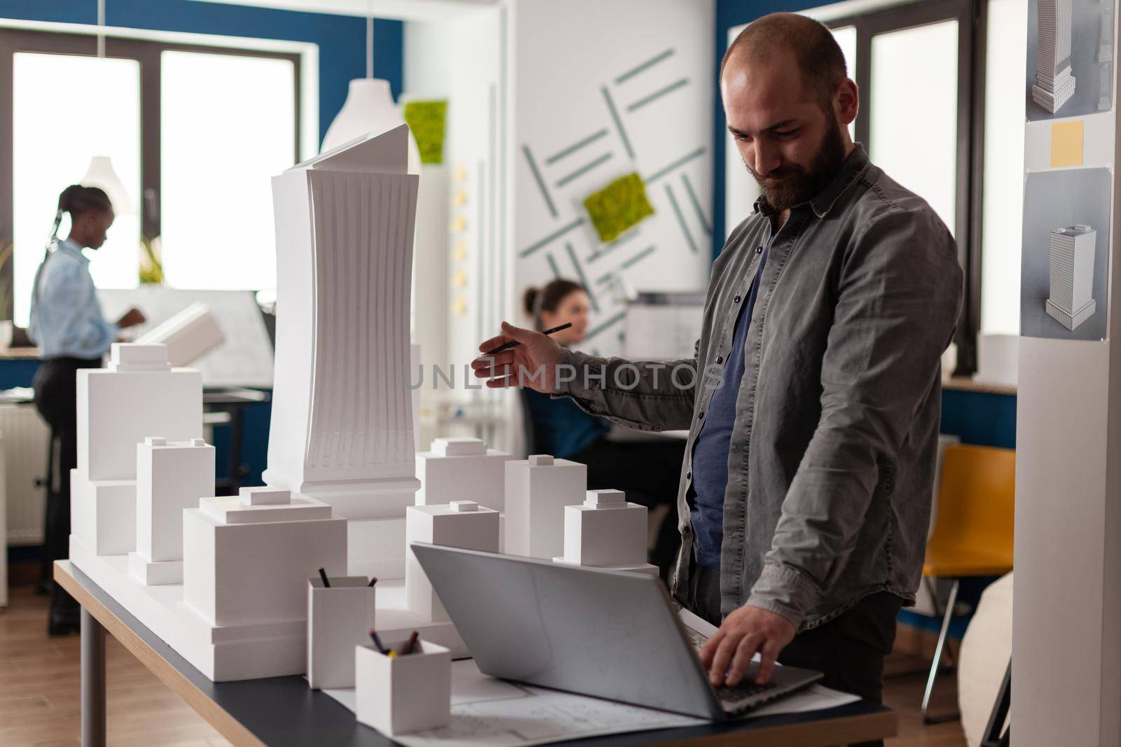Arhitectural designer in videocall conference presenting white foam building model of residential buildings to client. Architect using laptop computer in modern office next to desk with 3d maquette.