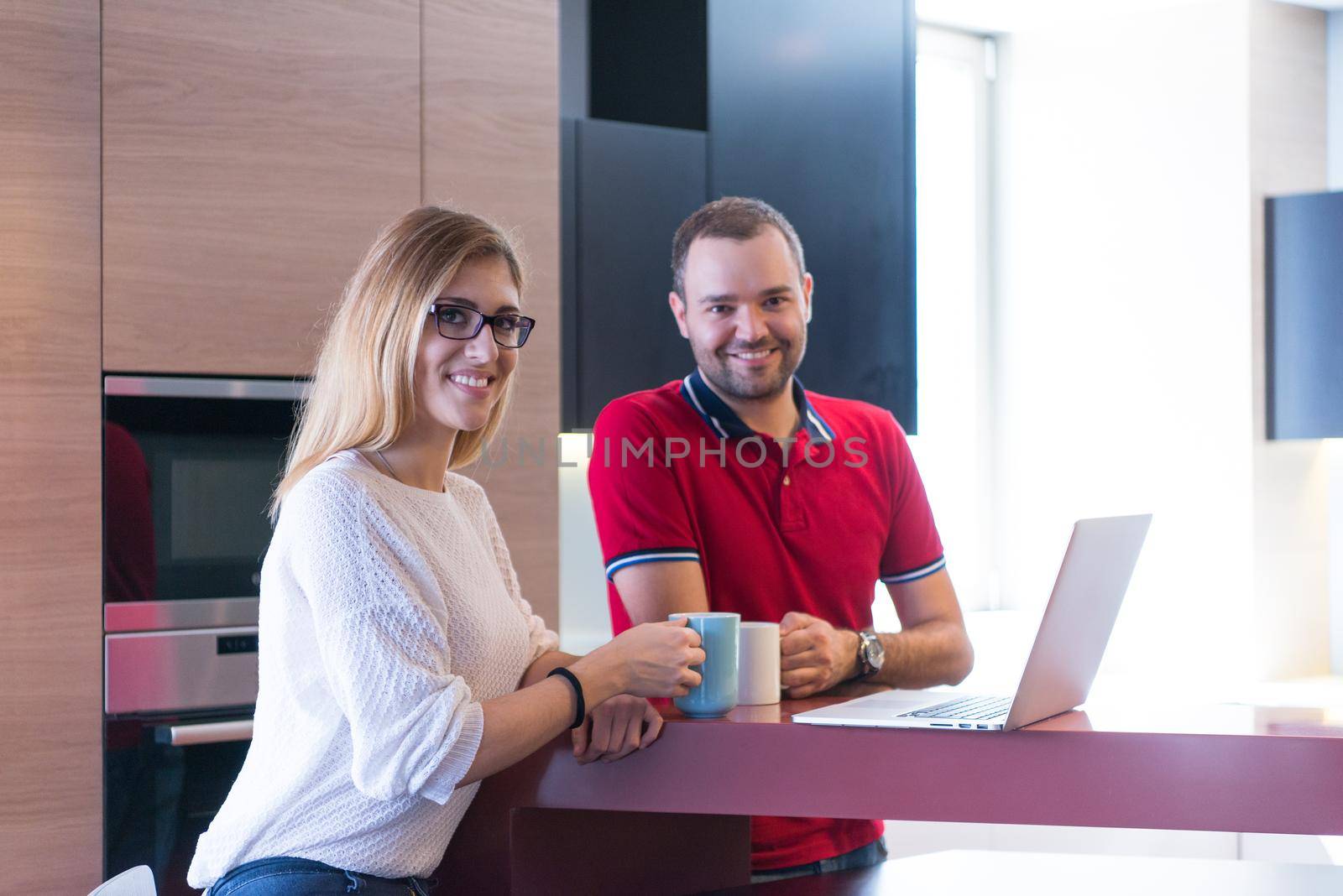 Young couple drinking coffee and using laptop computer at luxury home together, looking at screen, smiling.