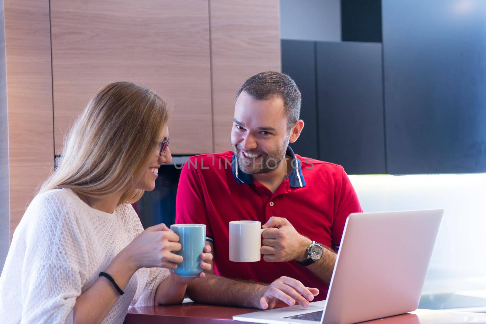 Young couple drinking coffee and using laptop computer at luxury home together, looking at screen, smiling.