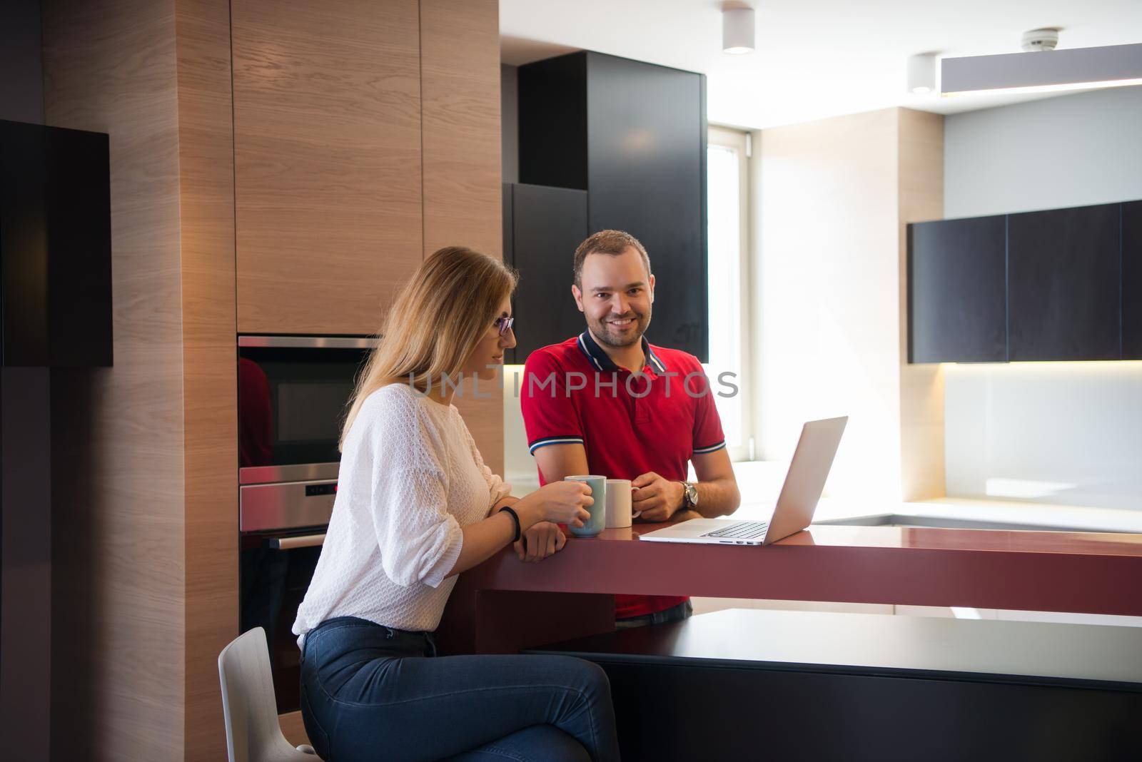 Young couple drinking coffee and using laptop computer at luxury home together, looking at screen, smiling.