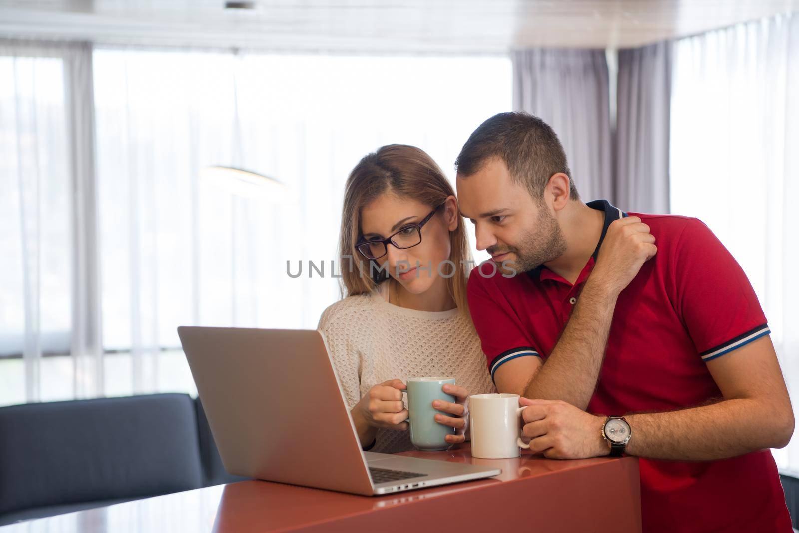 Young couple drinking coffee and using laptop computer at luxury home together, looking at screen, smiling.