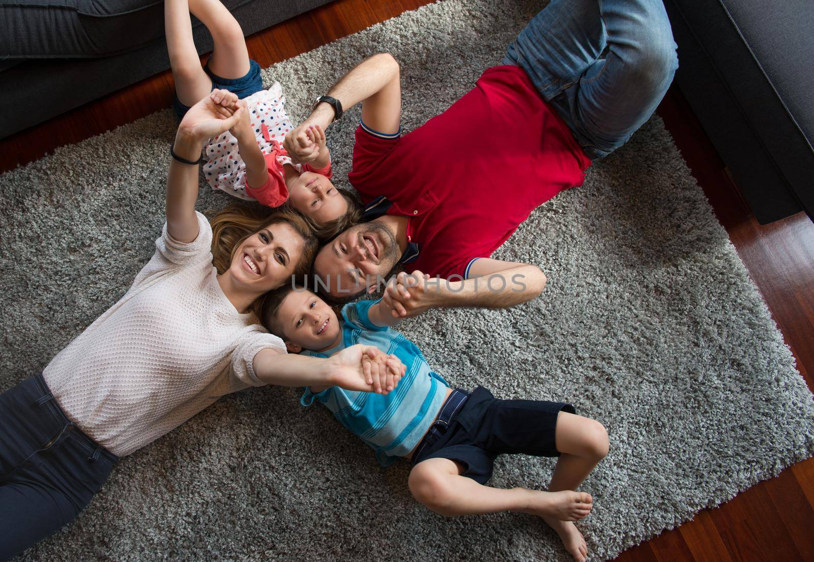 Top view of beautiful young parents, their cute little daughter and son holding hands, looking at camera and smiling, lying on the floor