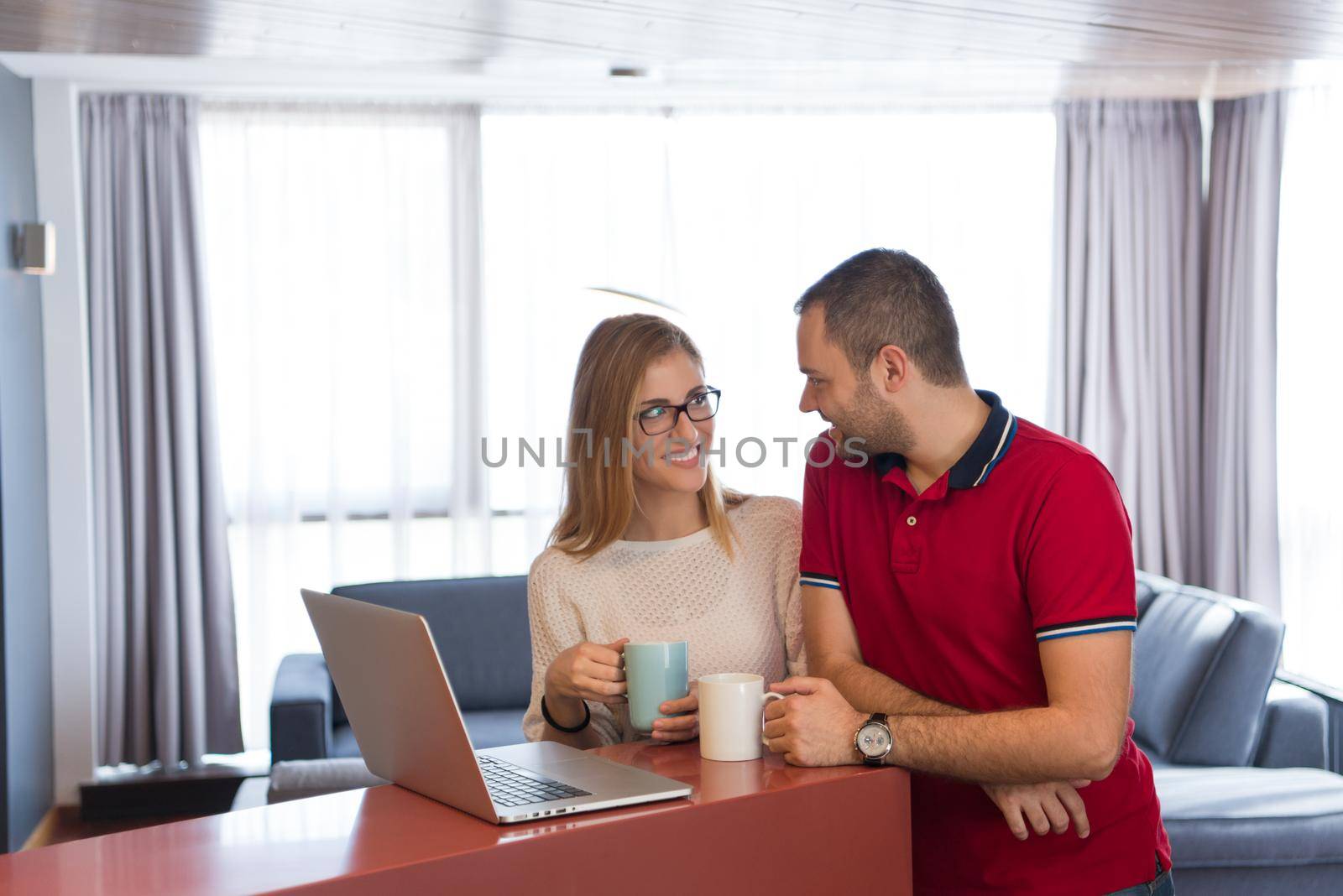 Young couple drinking coffee and using laptop computer at luxury home together, looking at screen, smiling.