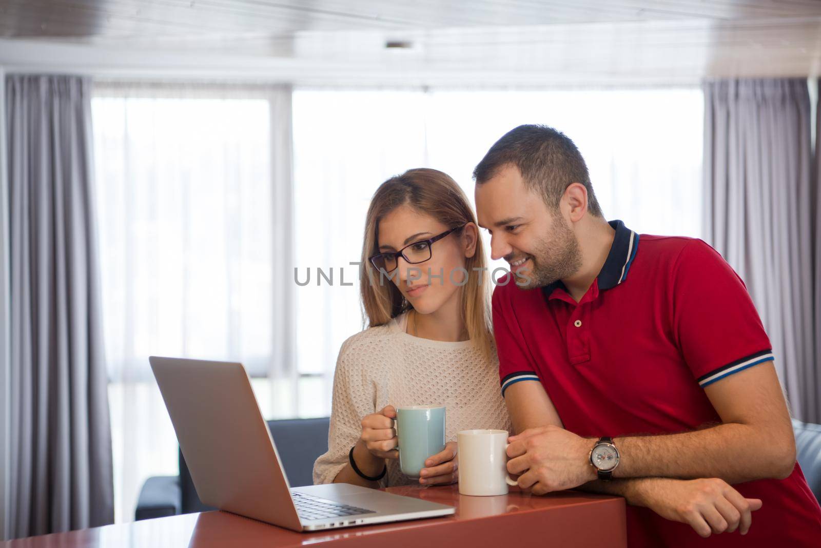 Young couple drinking coffee and using laptop computer at luxury home together, looking at screen, smiling.