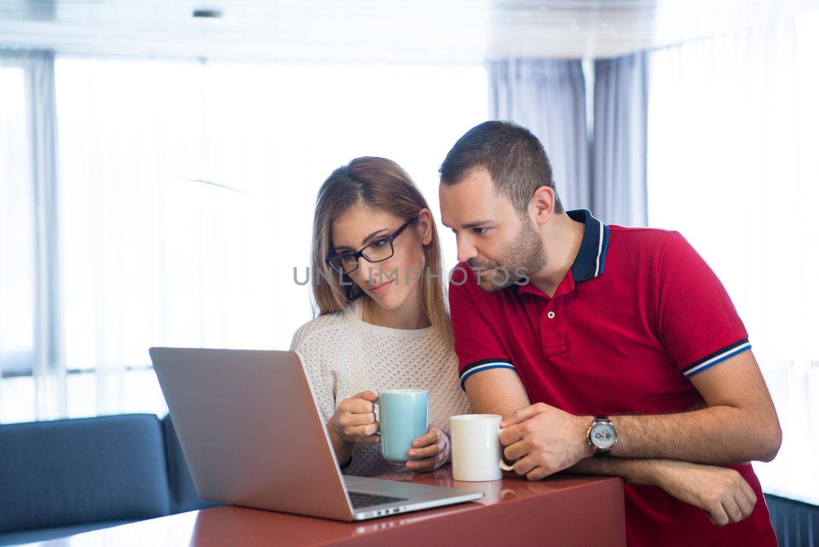 Young couple drinking coffee and using laptop computer at luxury home together, looking at screen, smiling.