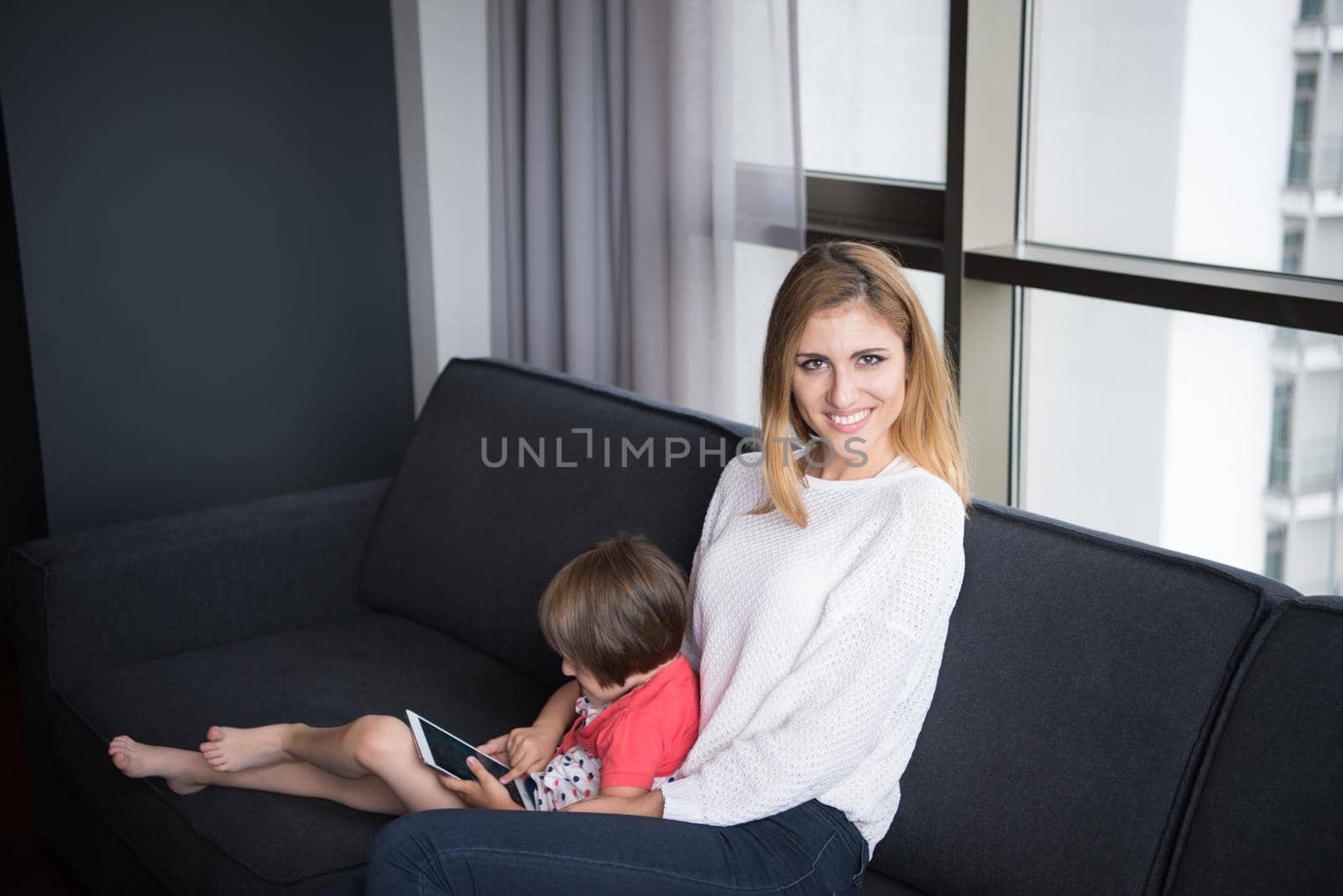 Beautiful young mother and her cute little daughter are using a tablet and smiling, sitting on sofa at home