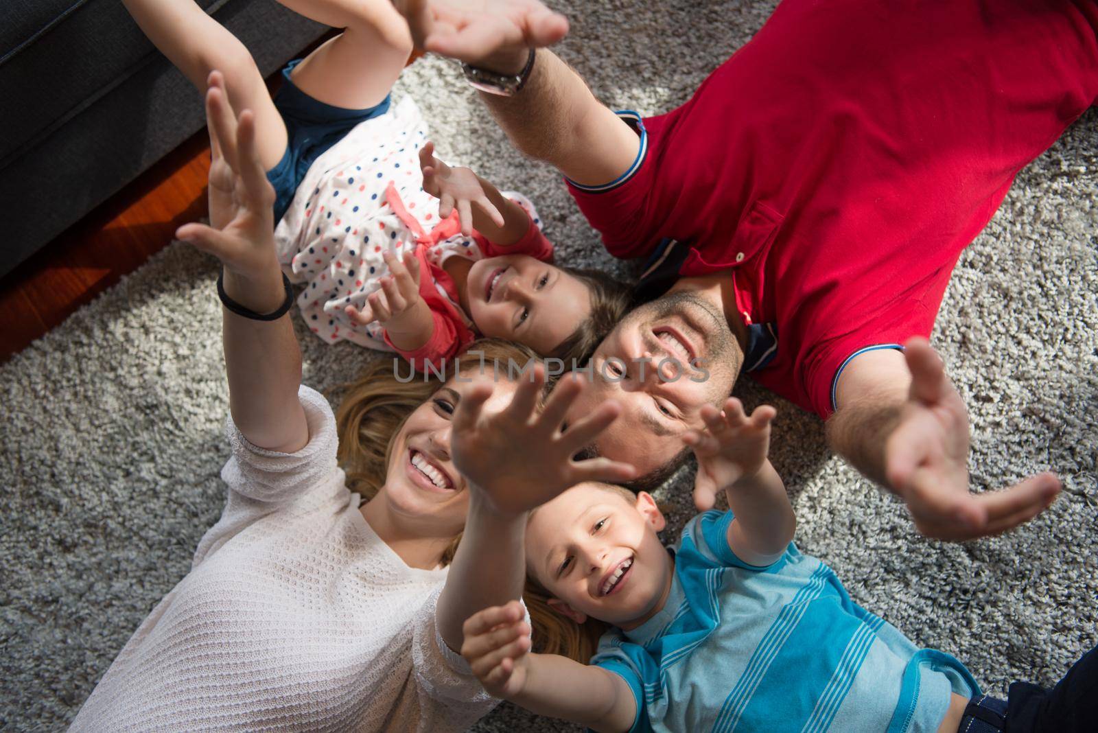 Top view of beautiful young parents, their cute little daughter and son holding hands, looking at camera and smiling, lying on the floor