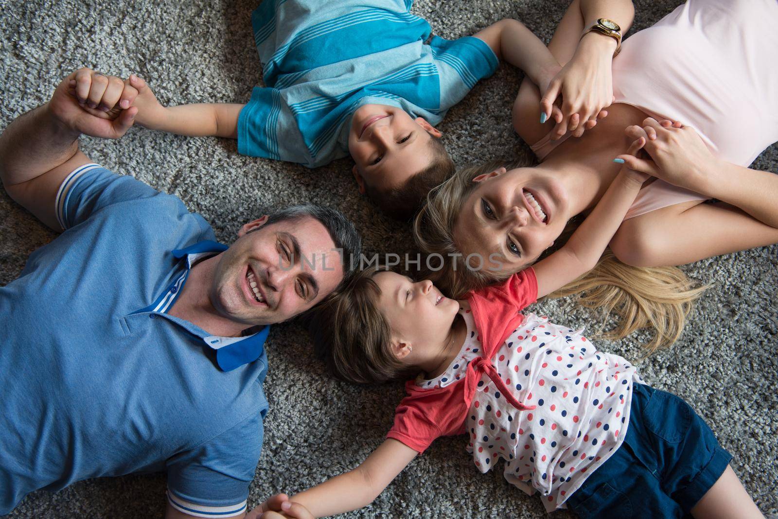 Top view of beautiful young parents, their cute little daughter and son holding hands, looking at camera and smiling, lying on the floor